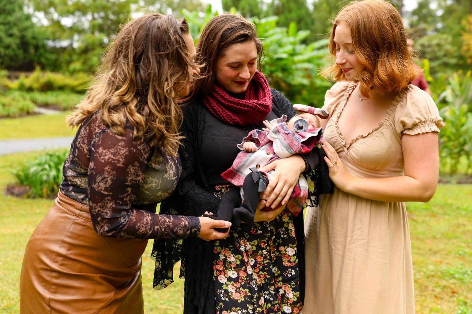 Three women in fall attire gather outdoors to admire a baby.