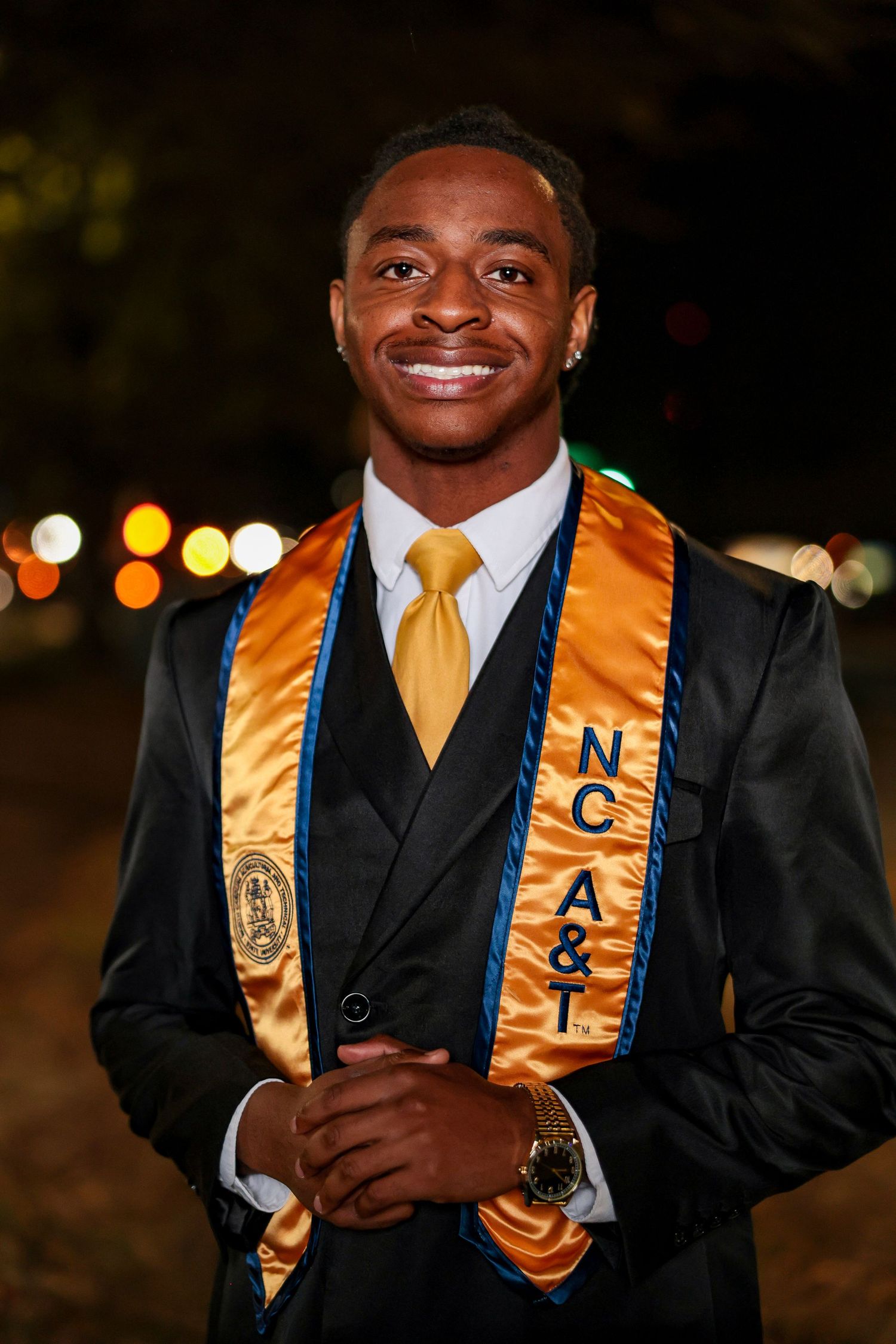 Graduate in black suit with gold stoles poses for nighttime graduation photos.