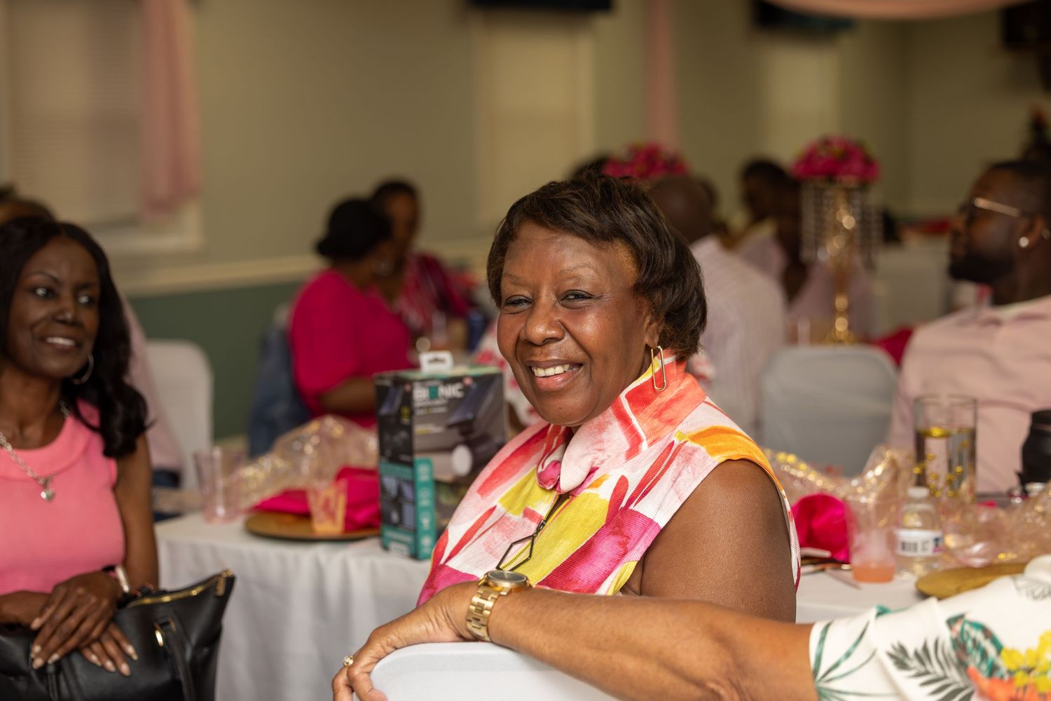 A joyful woman in colorful attire smiles at a festive pink-themed banquet event surrounded by guests.