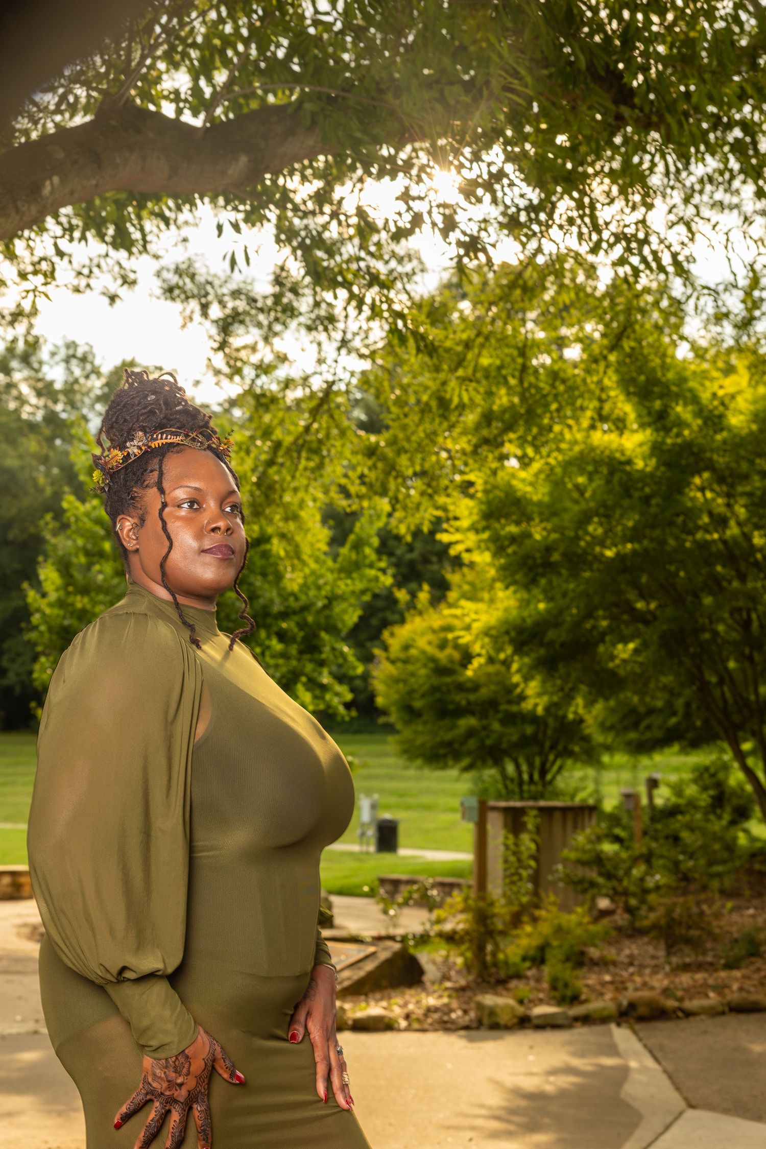 Woman in olive green dress posing outdoors in a sunlit park with lush green trees in the background.