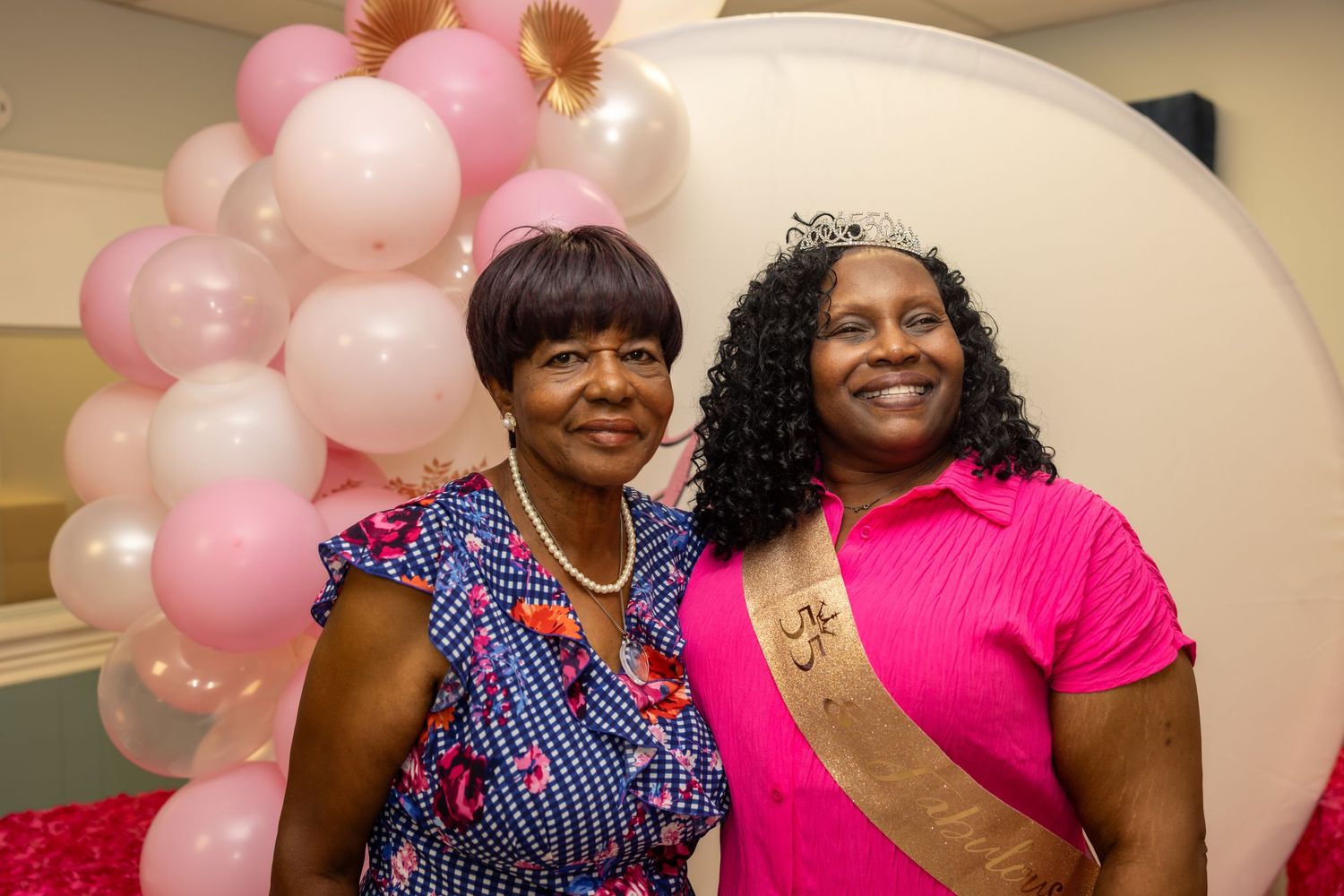 Two women pose together at a breast cancer awareness event with pink balloons, one wearing a tiara and sash.