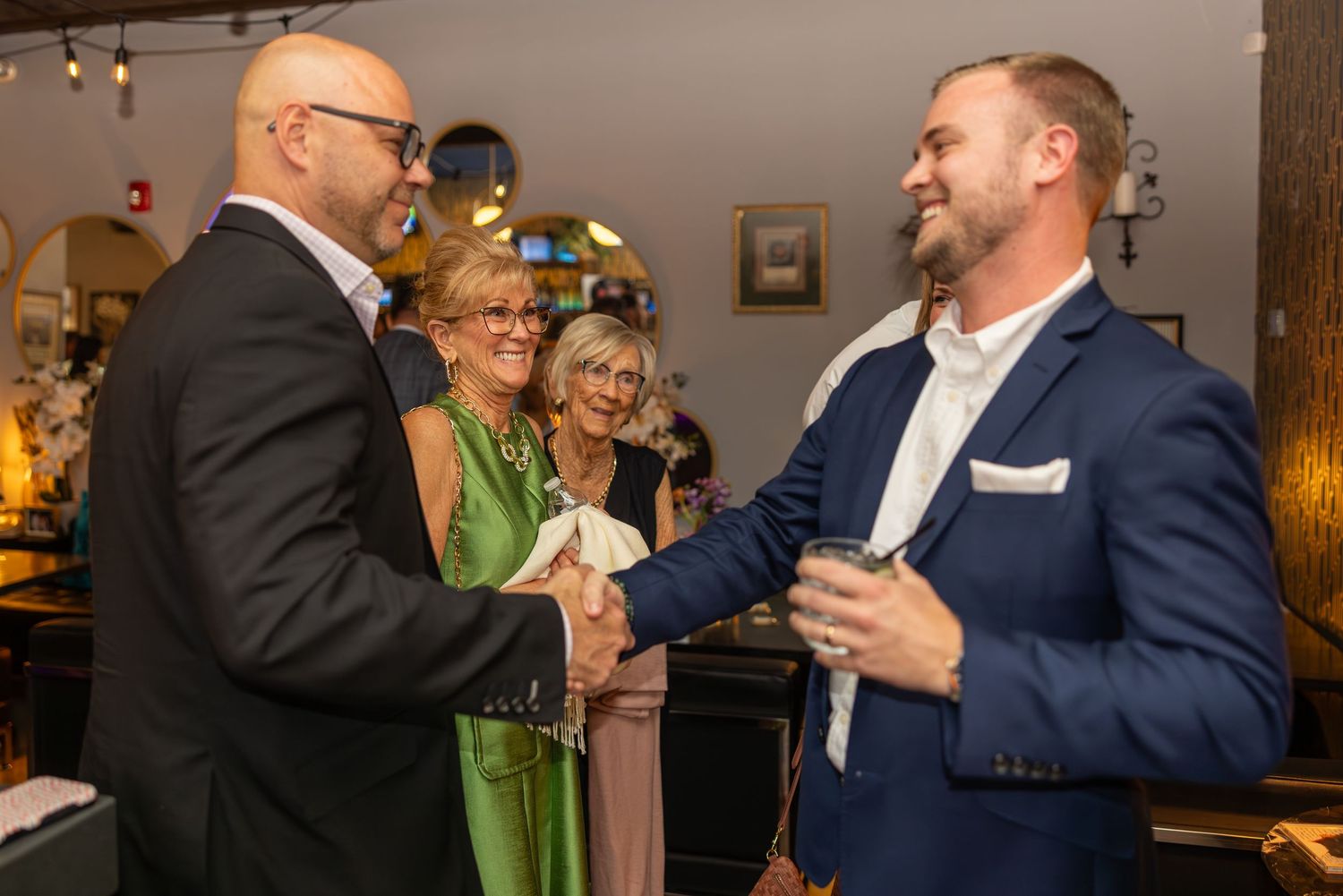 Two men shaking hands at an indoor networking event while two women smile in the background.