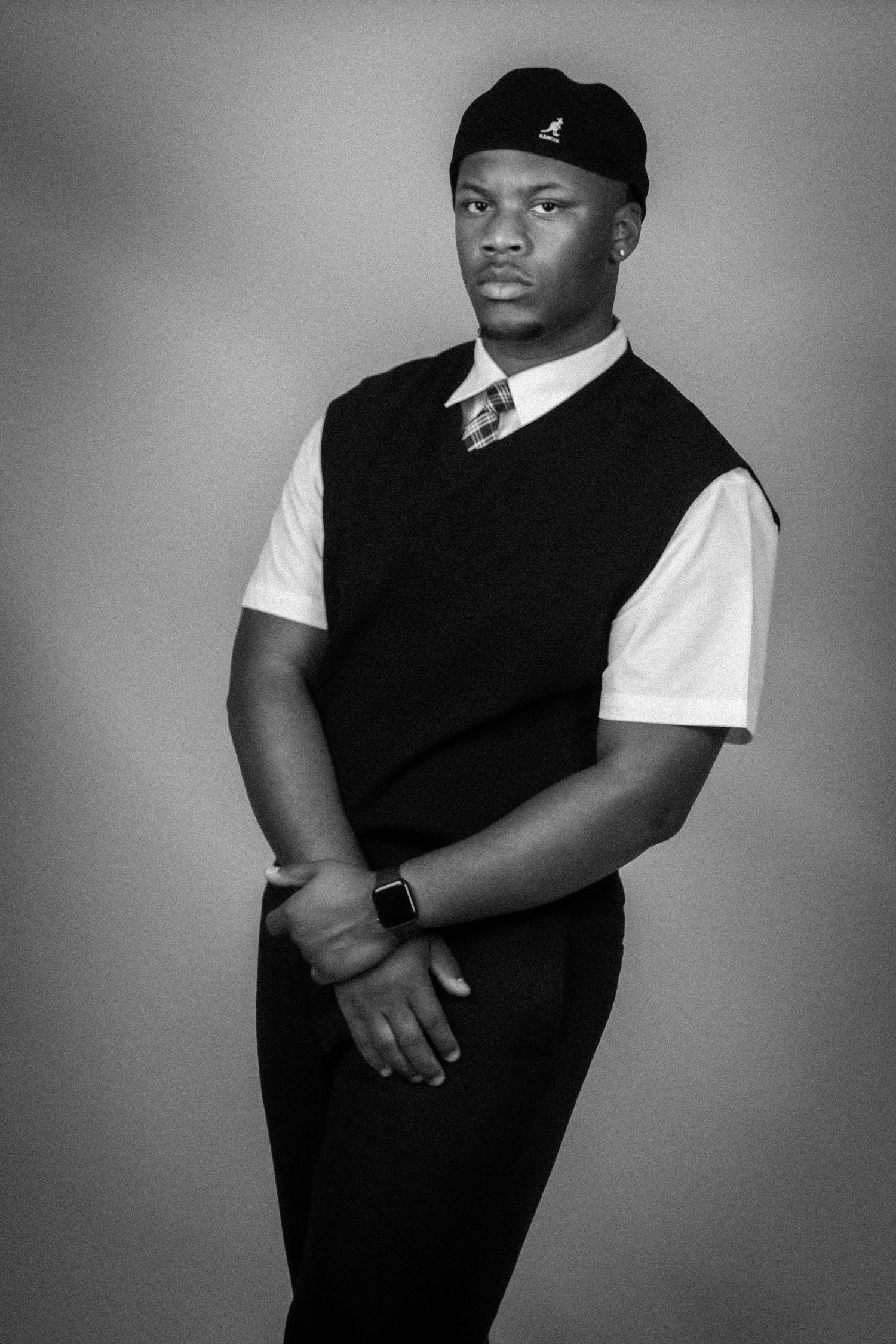 Black and white portrait of a young man in a black vest, white shirt, tie, and cap, posing confidently.