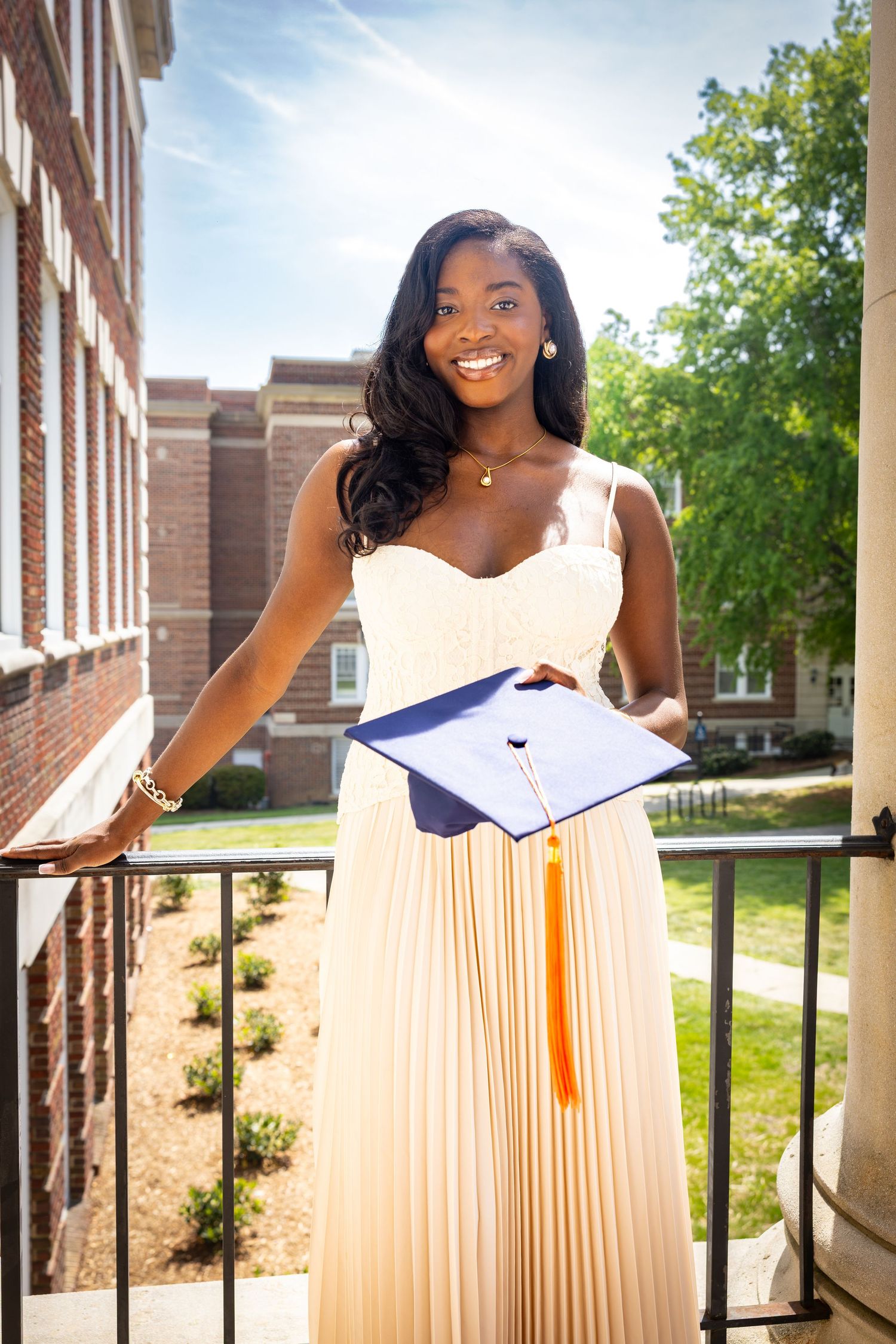 Smiling young woman in white dress holding graduation cap with orange tassel on university campus balcony.