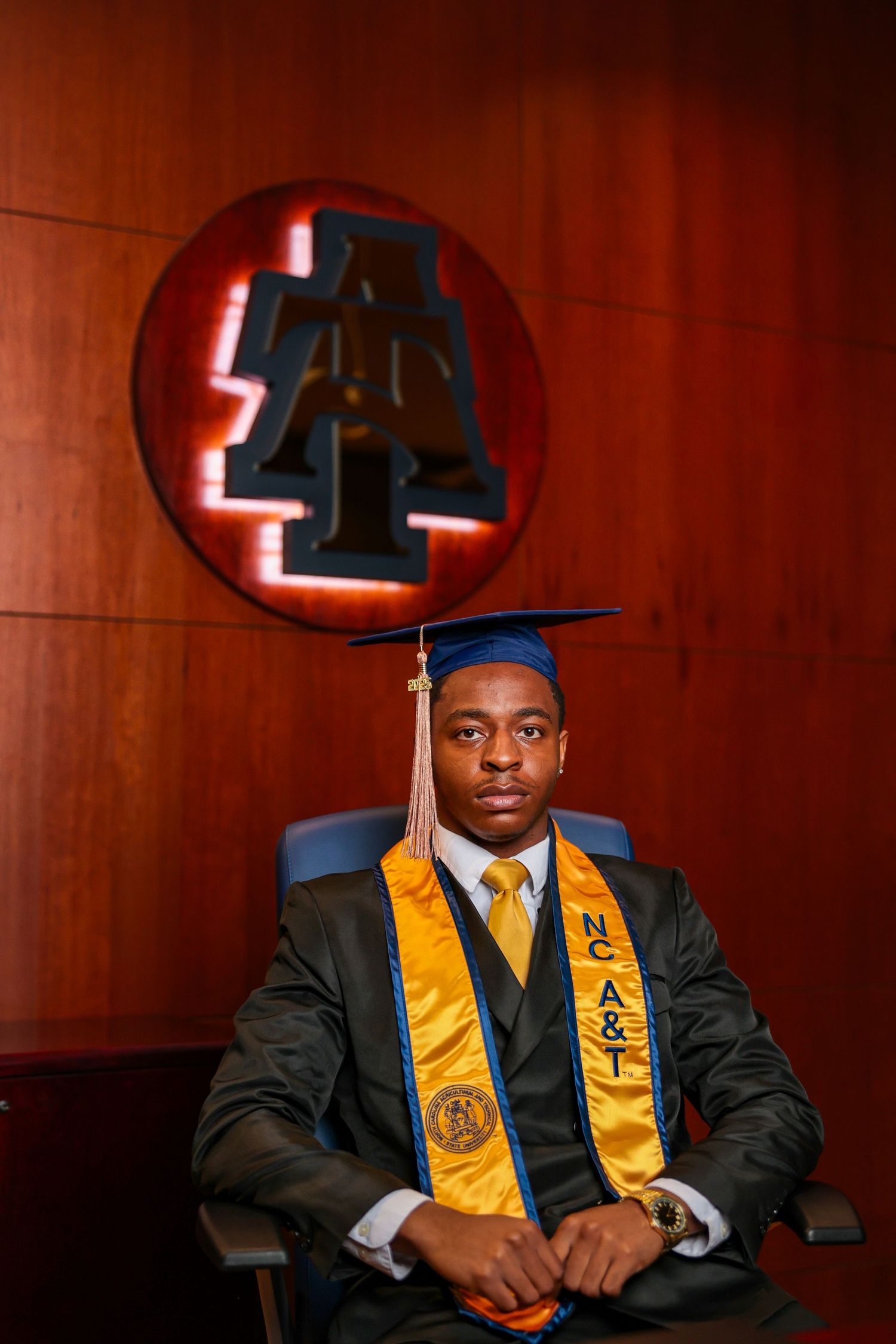Graduate wearing academic regalia sits in formal setting with wood paneling and decorative artwork.