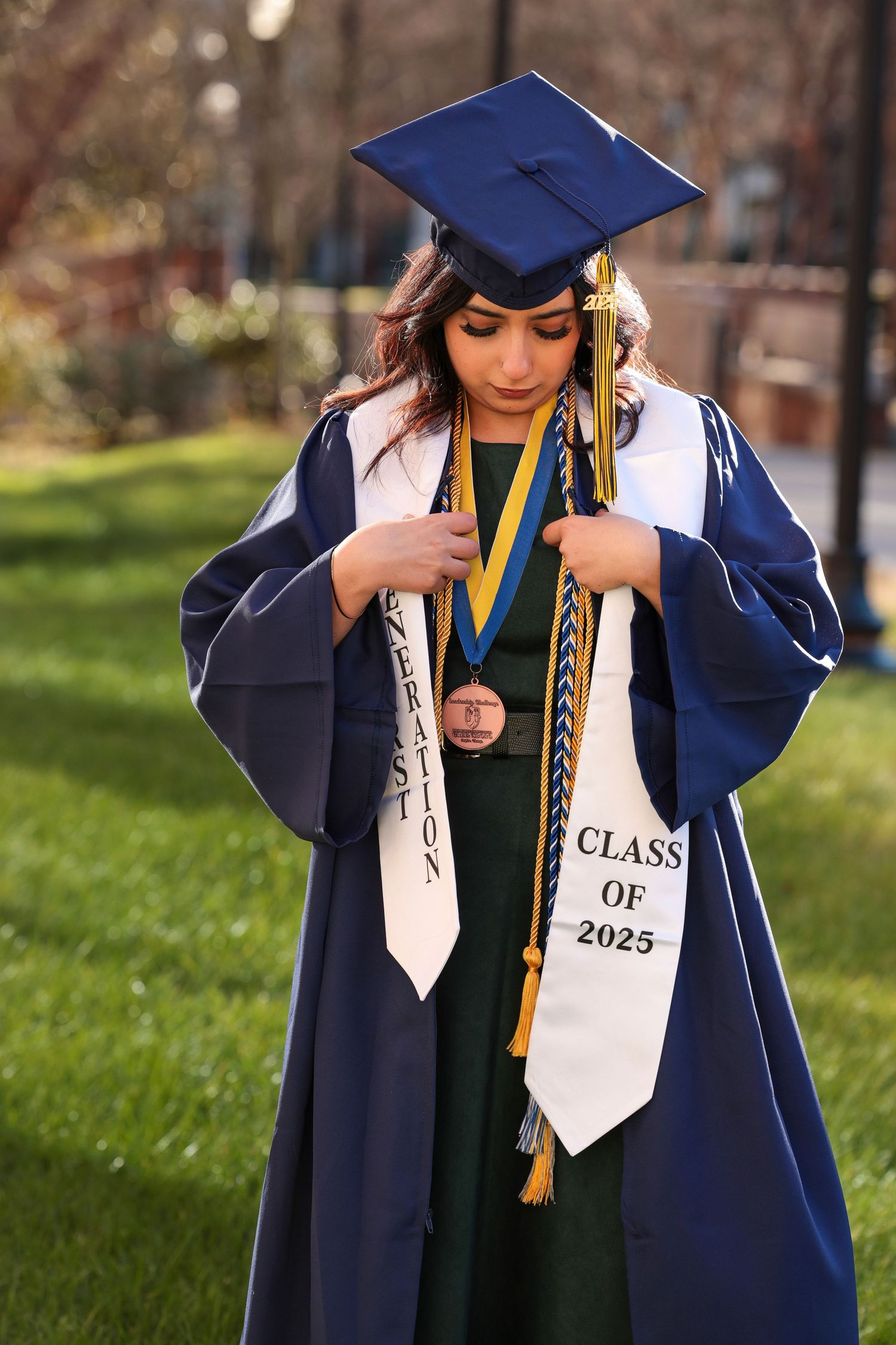 Graduate in navy blue cap and gown poses with Class of 2023 sash and graduation stoles on grassy campus.