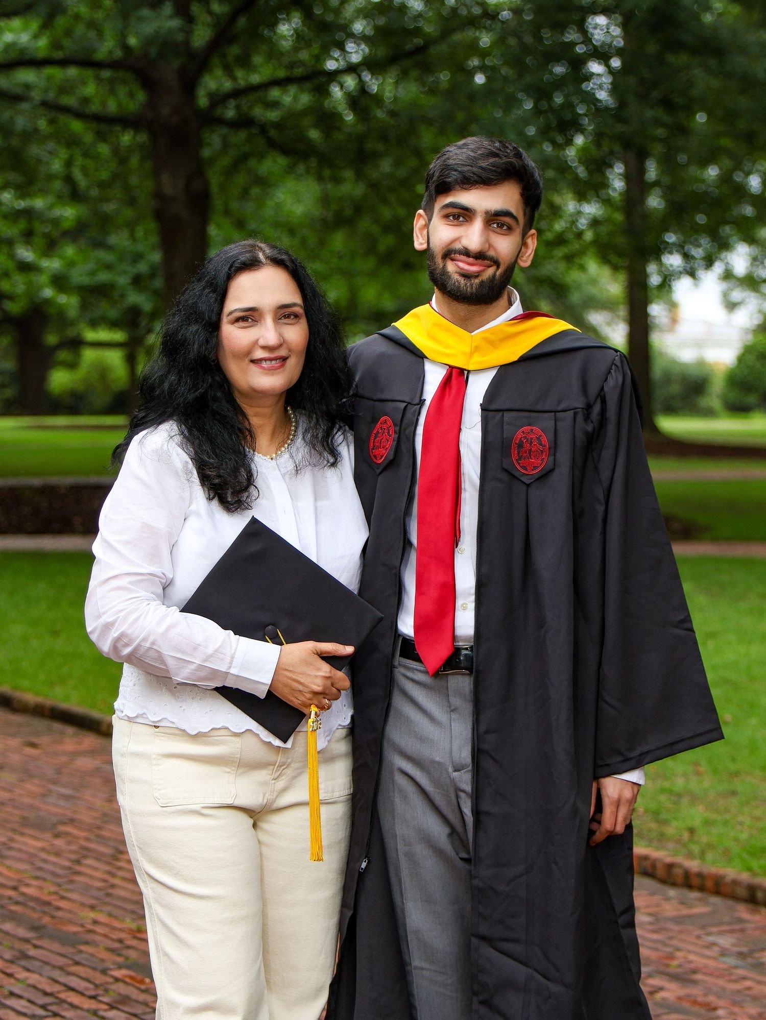 A graduate in academic regalia stands with a family member on a brick pathway.