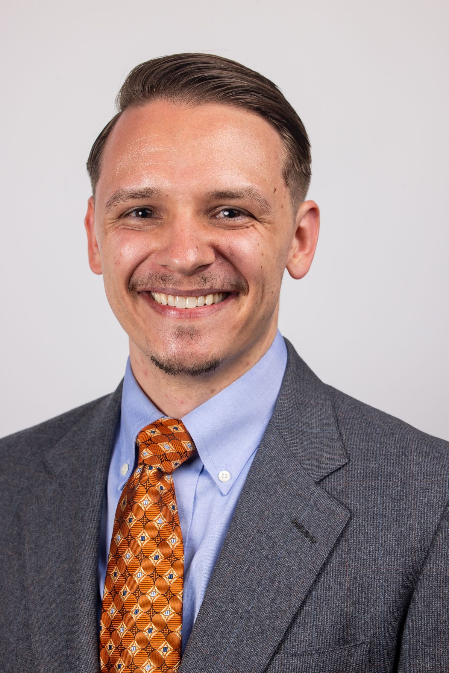 Professional headshot of a smiling man in a gray suit with an orange patterned tie.