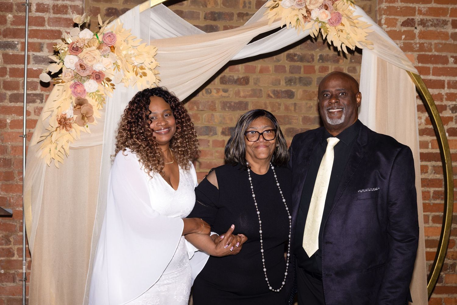 Three people posing under a floral arch with pink and white flowers at an elegant wedding ceremony venue.