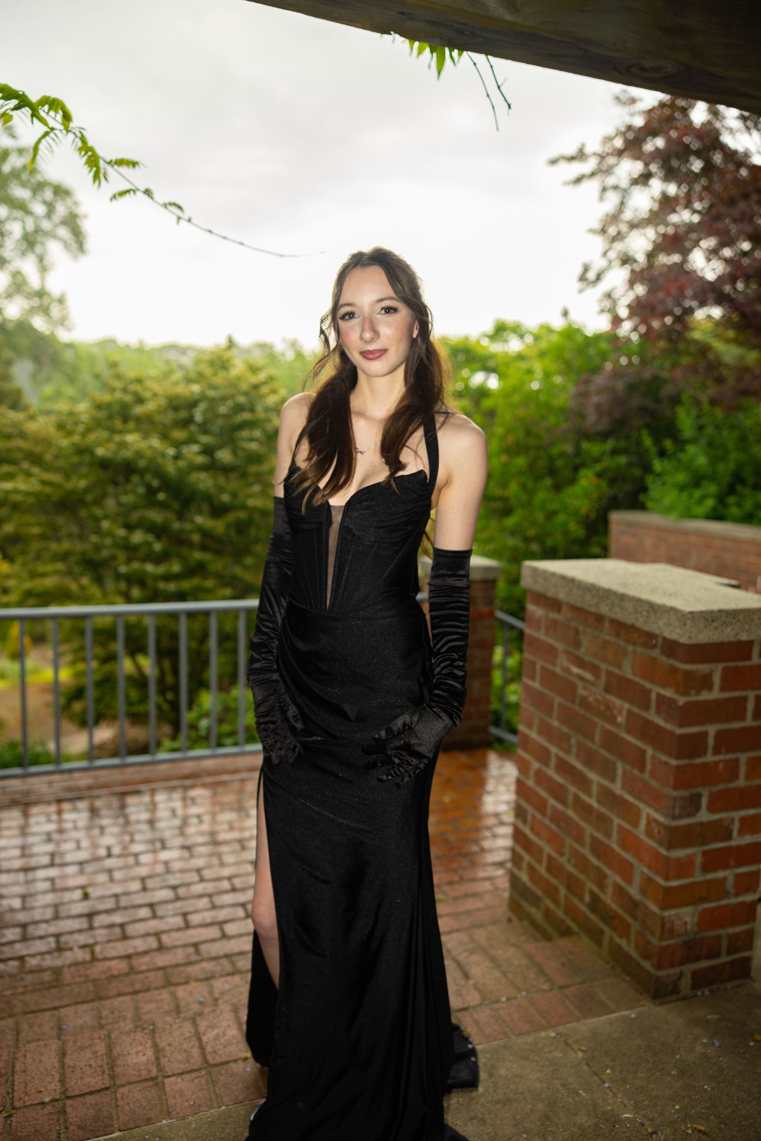 Young woman in elegant black off-shoulder gown with long gloves poses on brick terrace surrounded by lush greenery.
