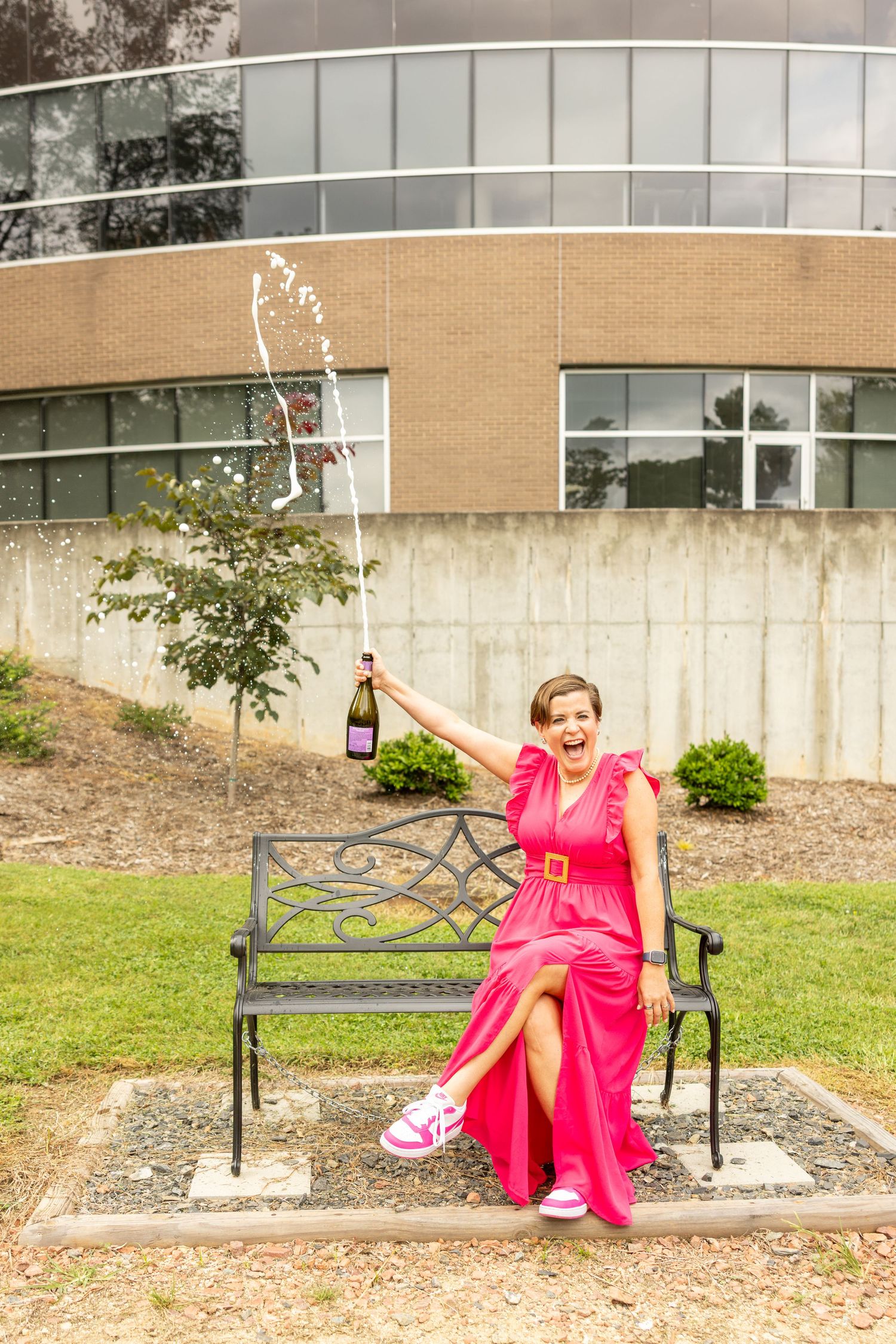 Woman in pink dress sitting on bench popping champagne bottle outdoors near modern brick building.