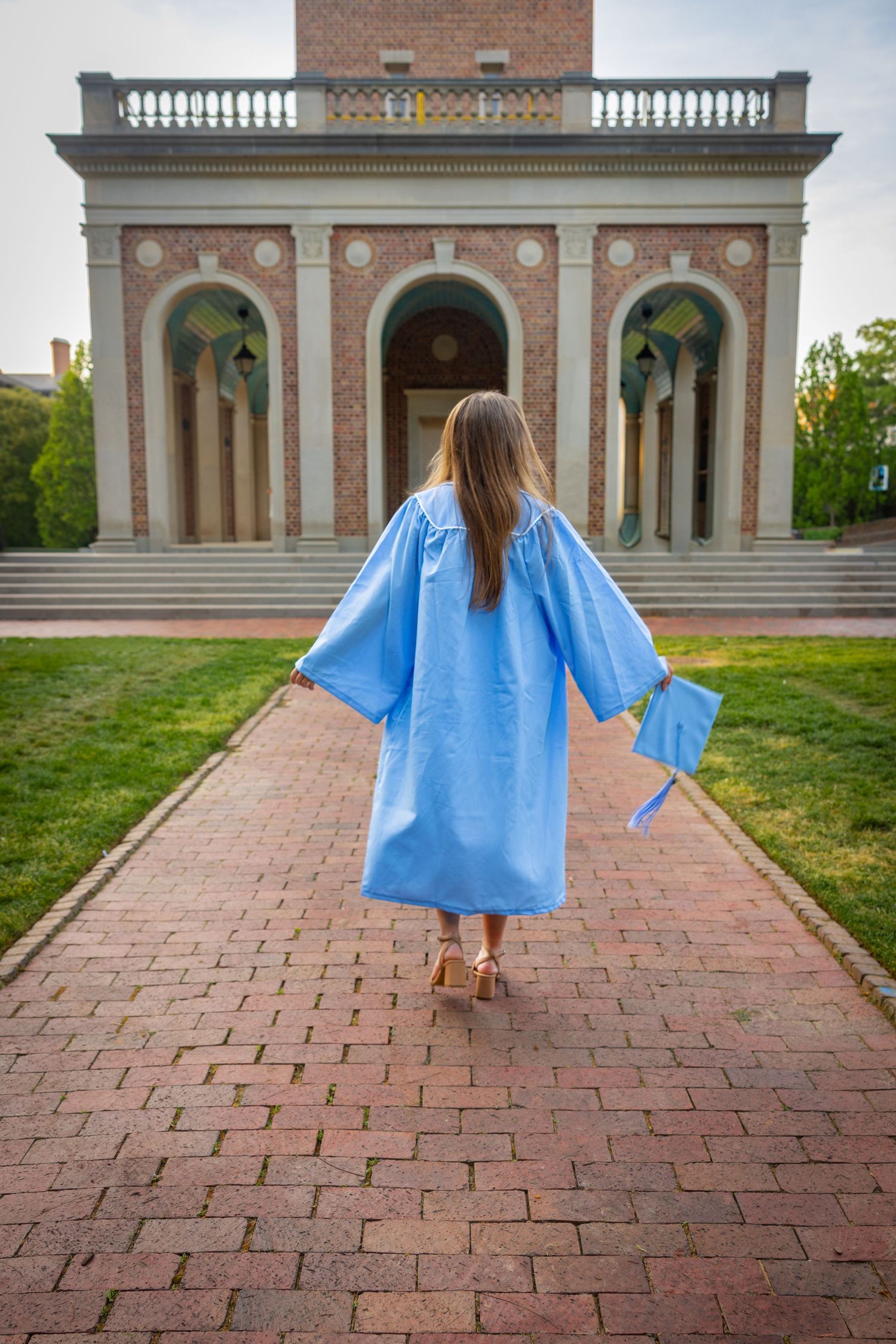 A graduate in a light blue cap and gown walks toward a brick building on a college campus.