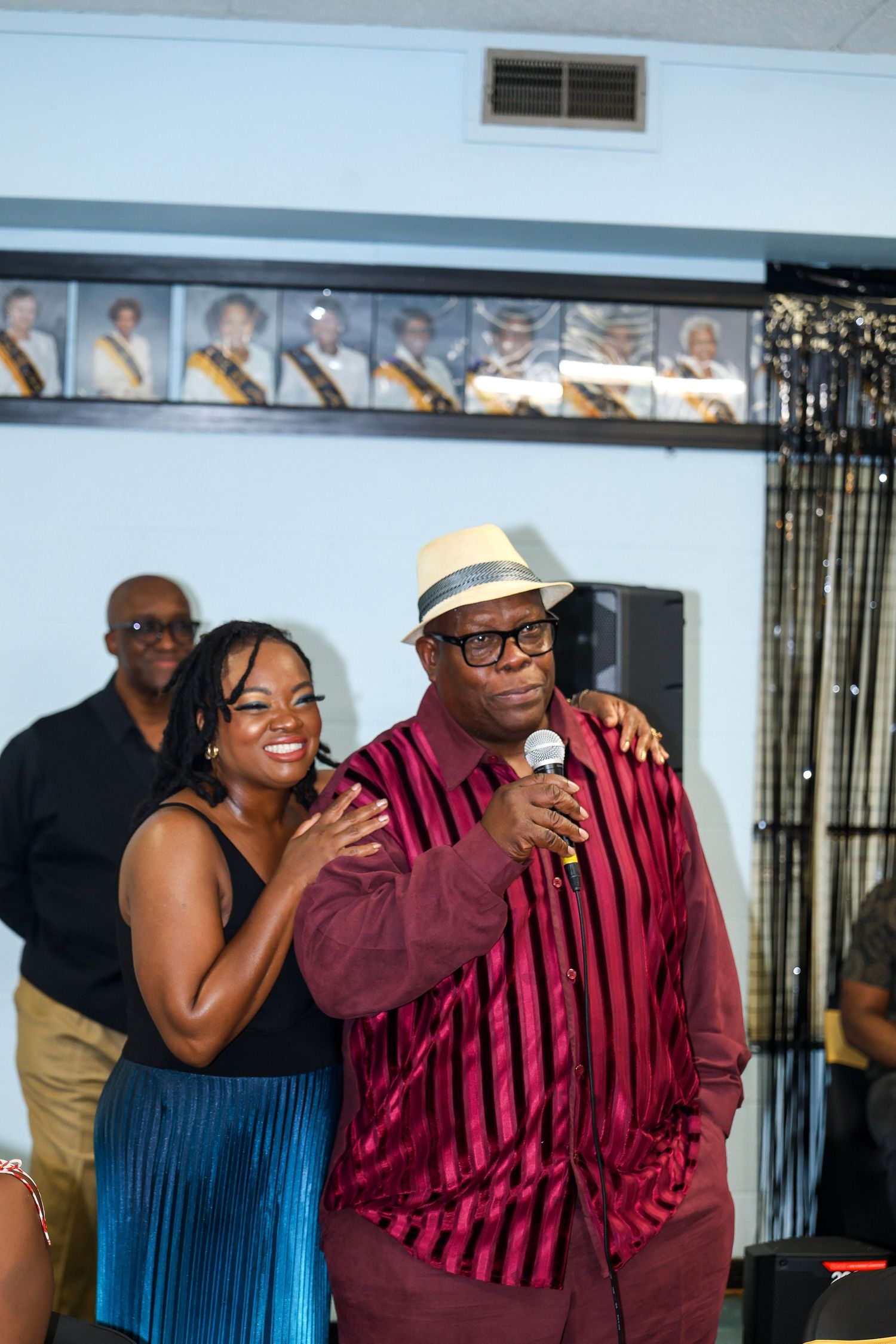 Two people singing and performing at a celebration with a row of photos visible on the wall behind them.