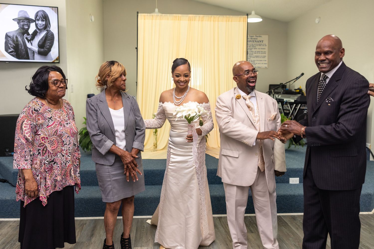 Bride in white gown stands with groom and guests smiling at a wedding ceremony inside a church.