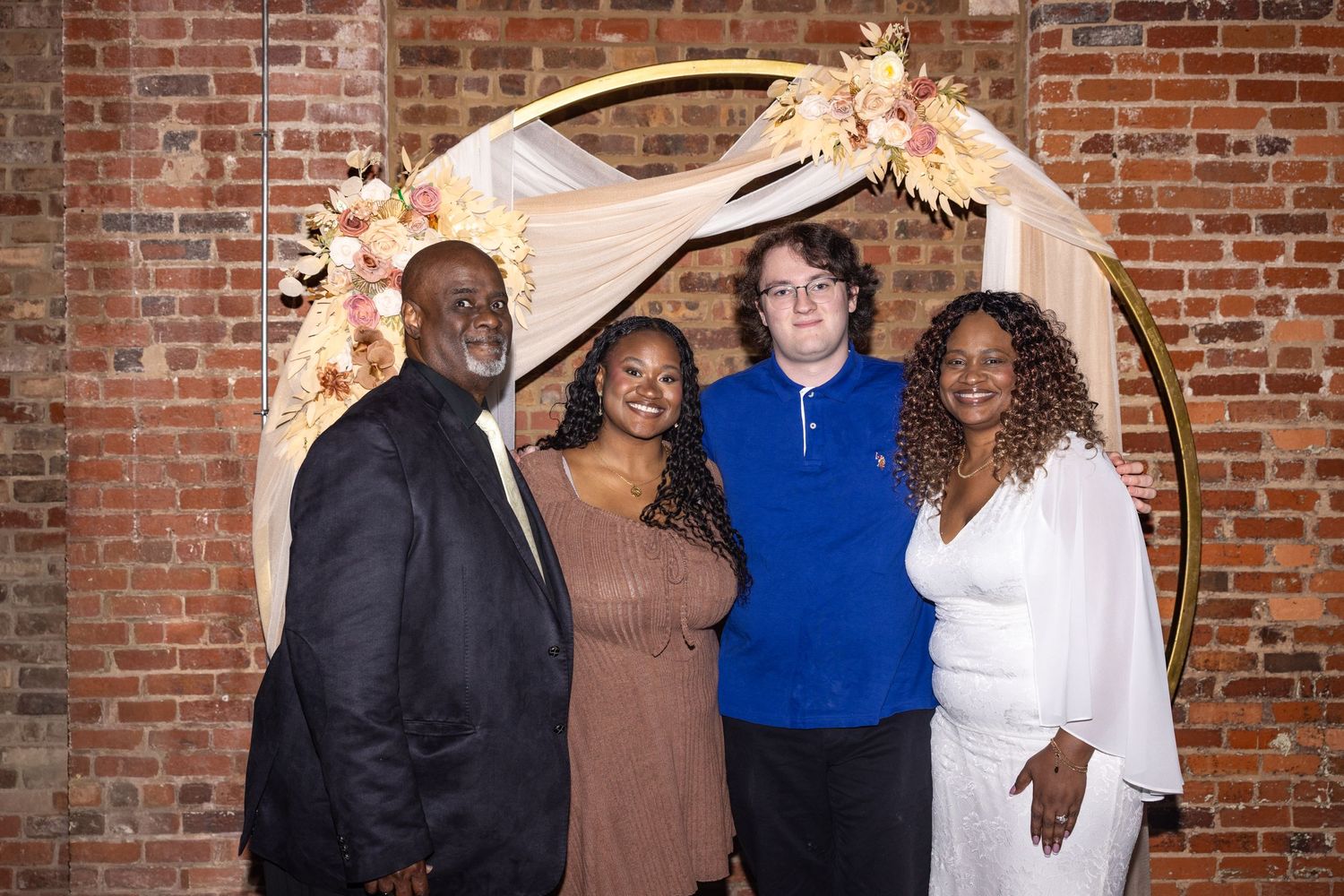Four people smiling together in front of a floral circular arch at a wedding venue with exposed brick walls.