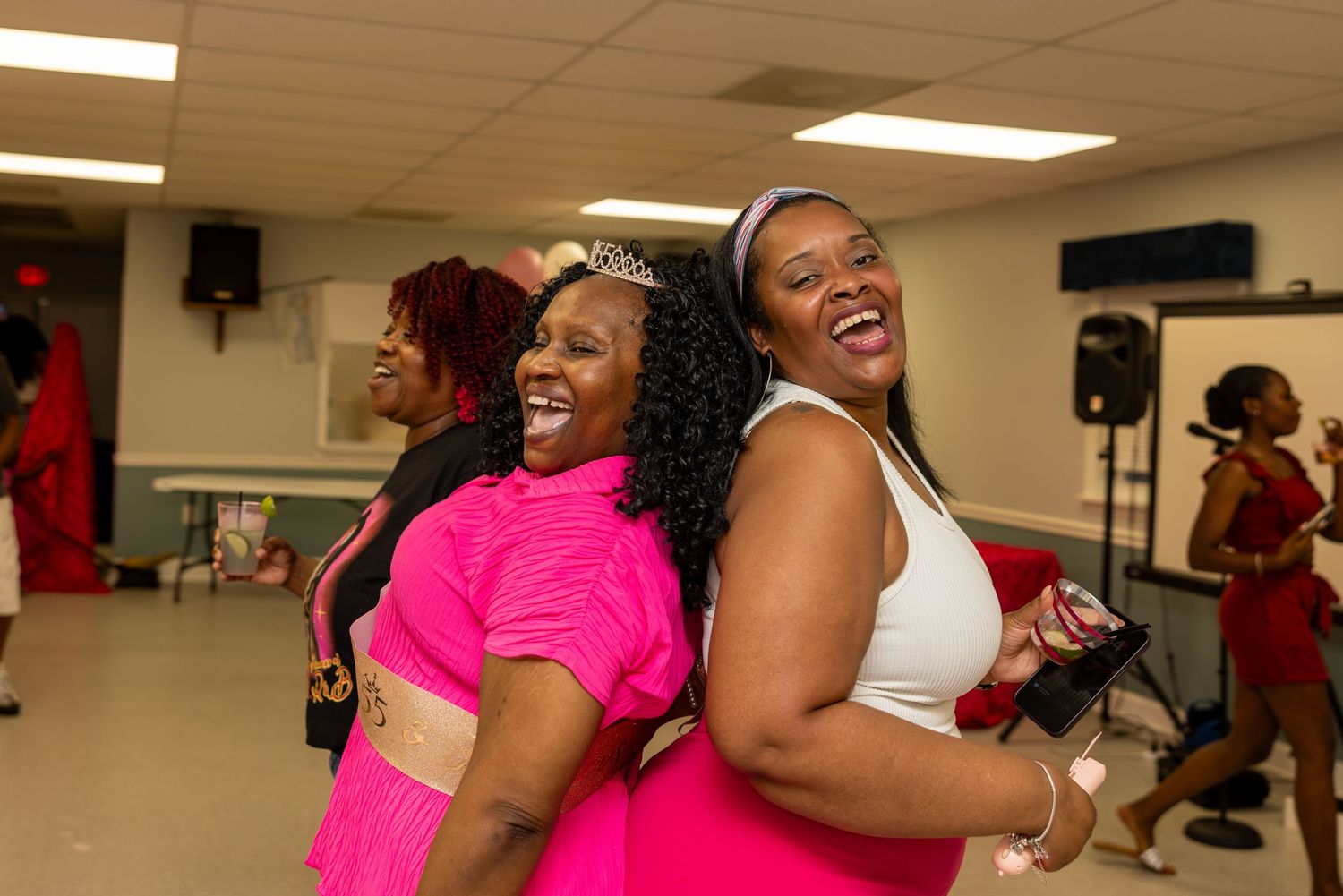 Two women laughing and dancing together at a festive party, one wearing a pink outfit and tiara.