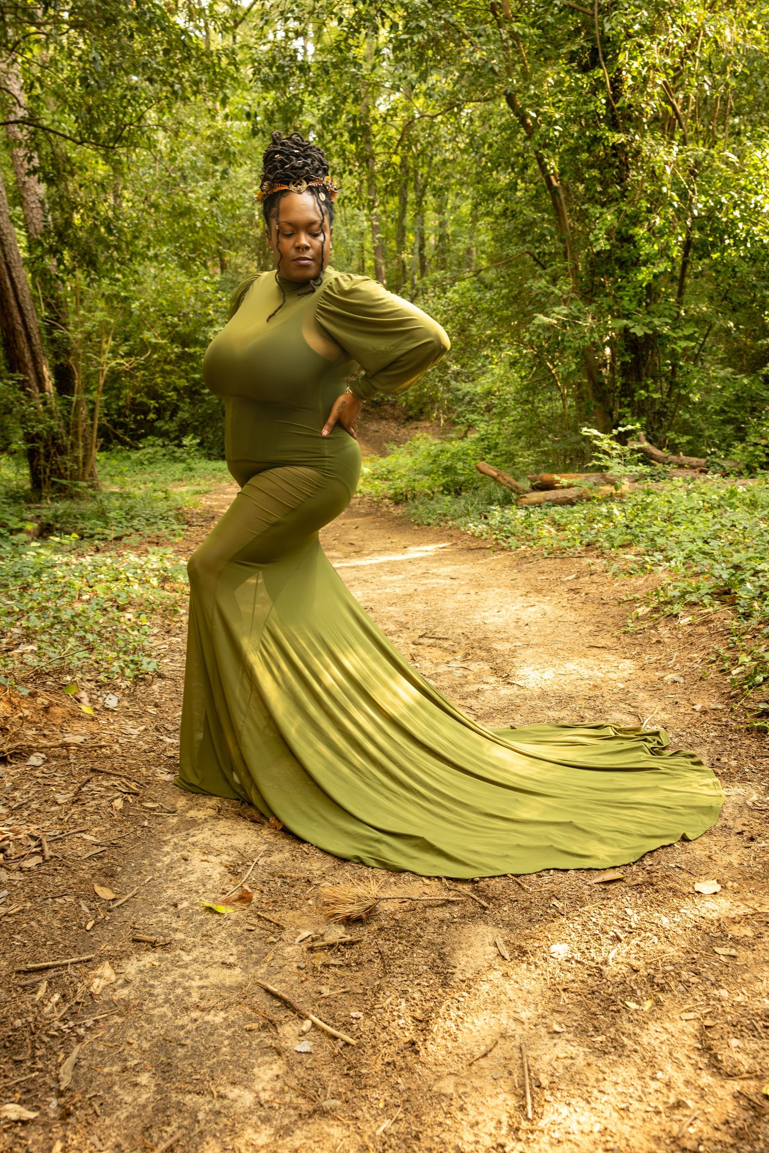 Woman in elegant olive green gown with train poses on forest path surrounded by lush green trees.