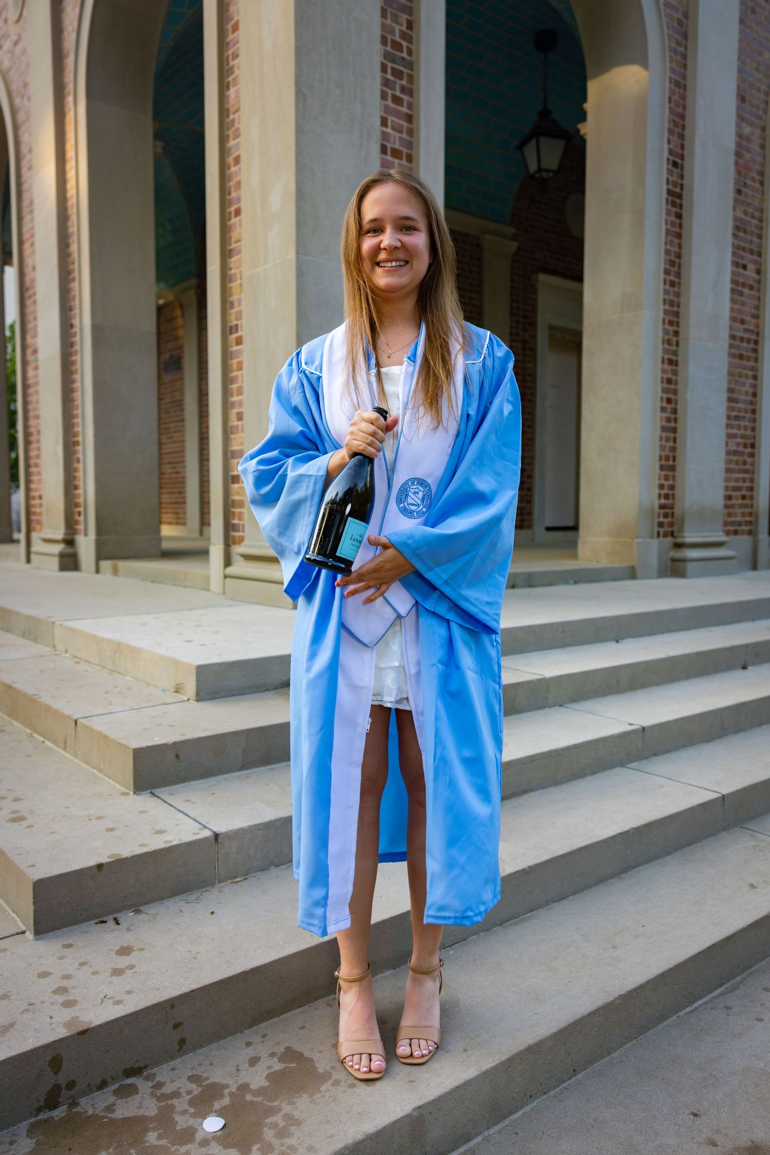 Smiling graduate in light blue graduation gown holding a bottle on university steps.