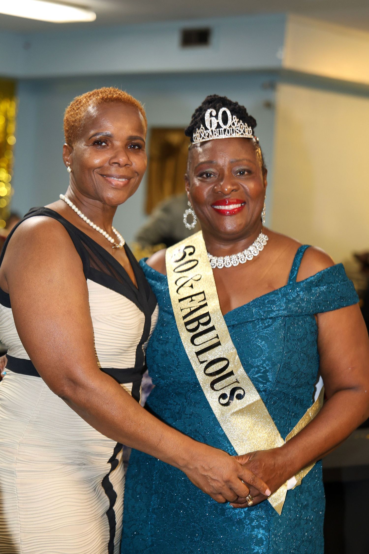 Two elegantly dressed women at a formal event wearing evening gowns and jewelry with one wearing a 60th birthday sash and tiara.