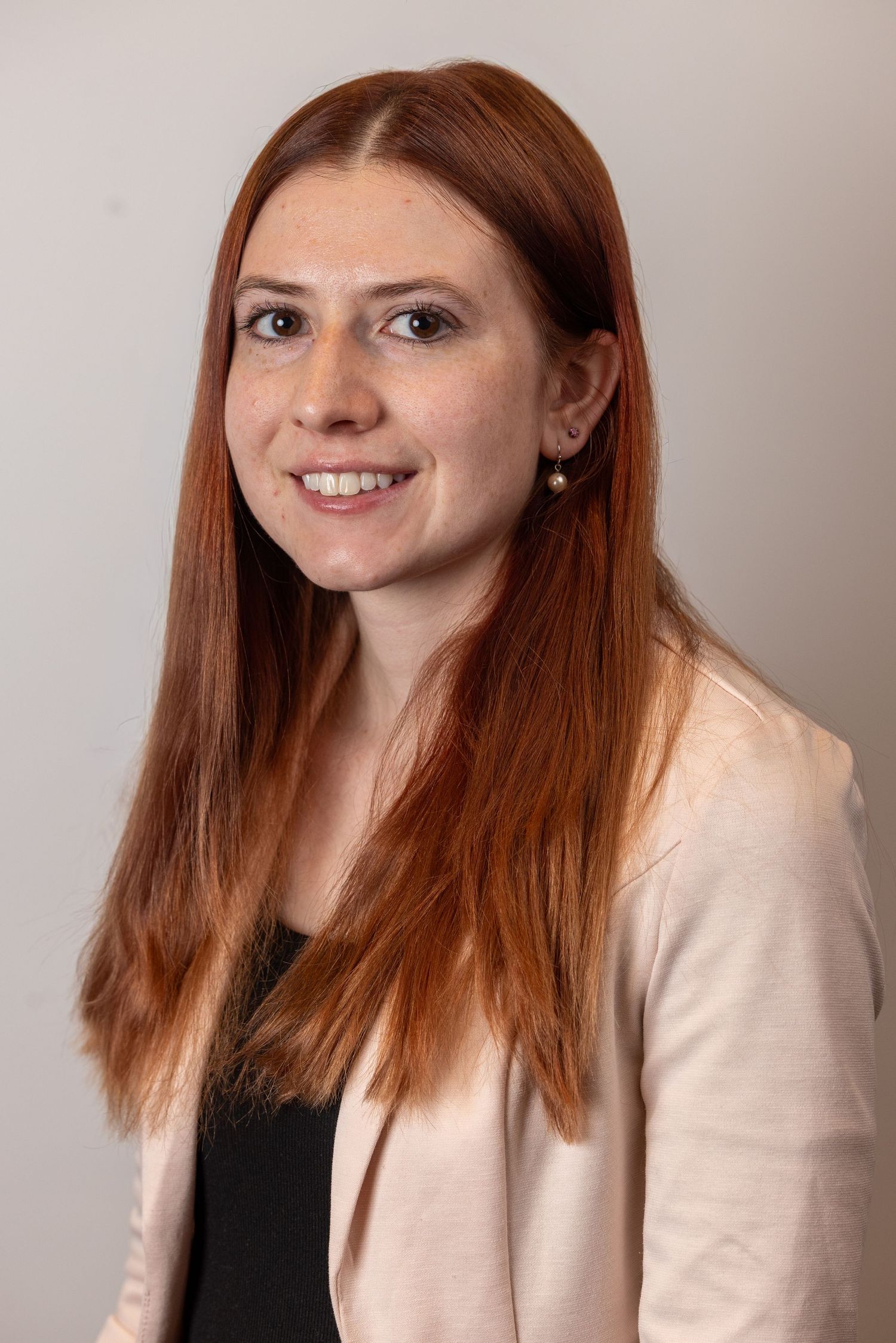 Professional headshot of a smiling young woman with long red hair wearing a light pink blazer.