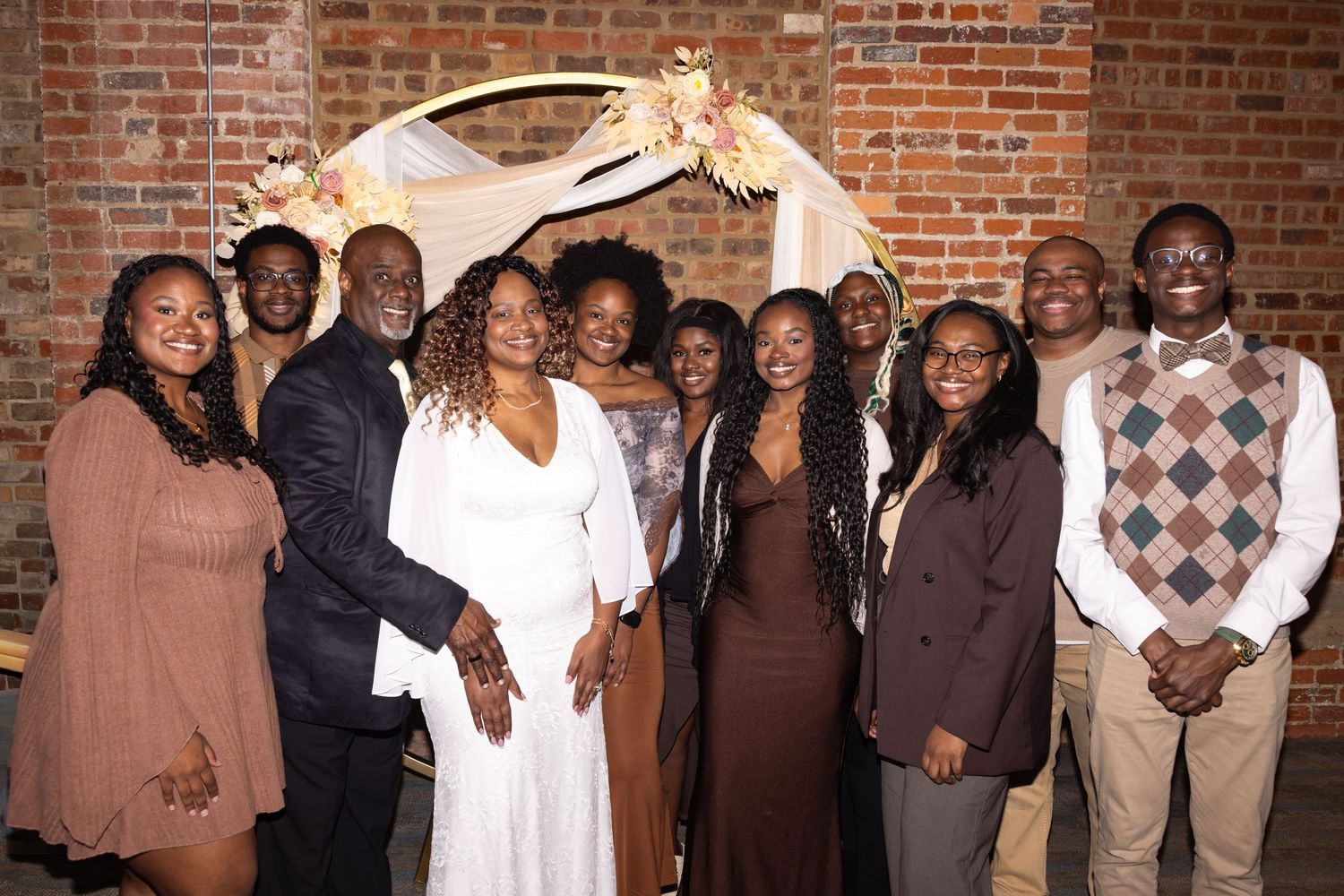 A joyful family group poses together under a floral arch at a wedding celebration in a brick-walled venue.