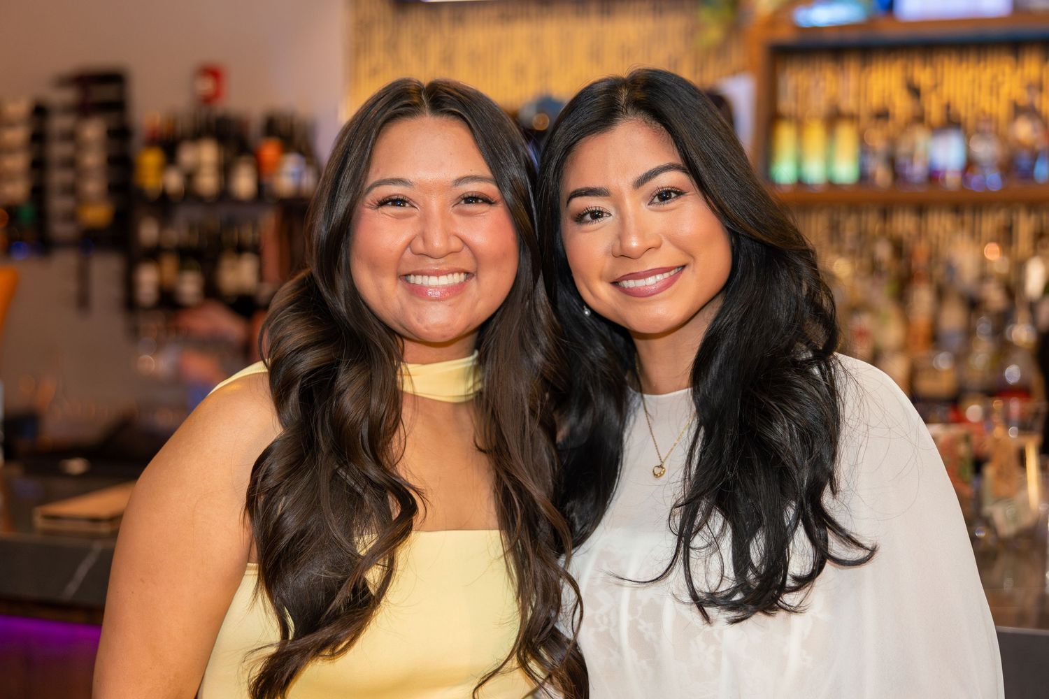 Two smiling young women with long dark hair pose together in front of a bar backdrop.