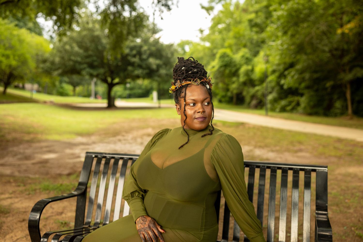 Woman in olive green outfit sitting on a park bench surrounded by lush green trees.