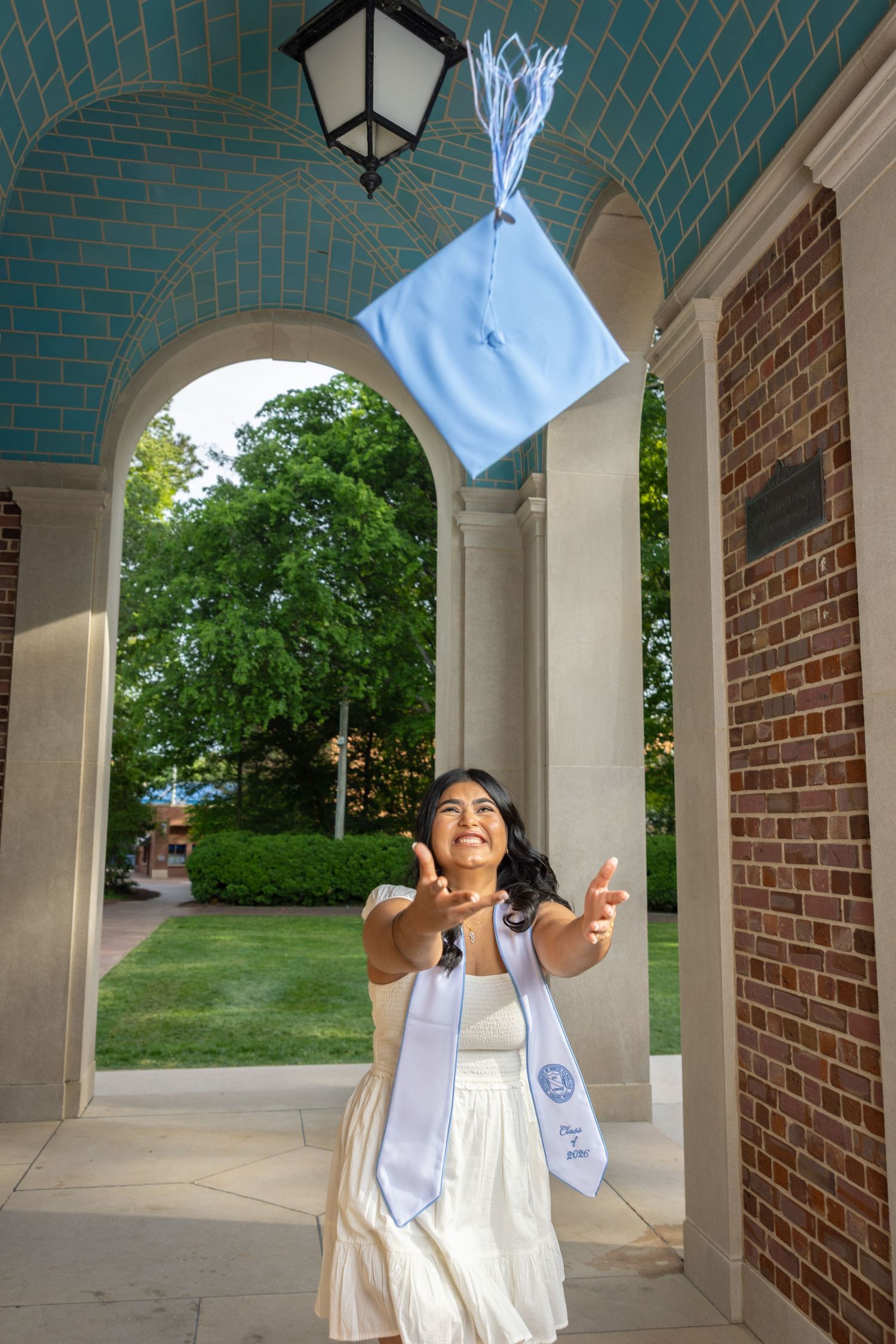Young woman in white dress tossing light blue graduation cap in the air under brick archway on college campus.