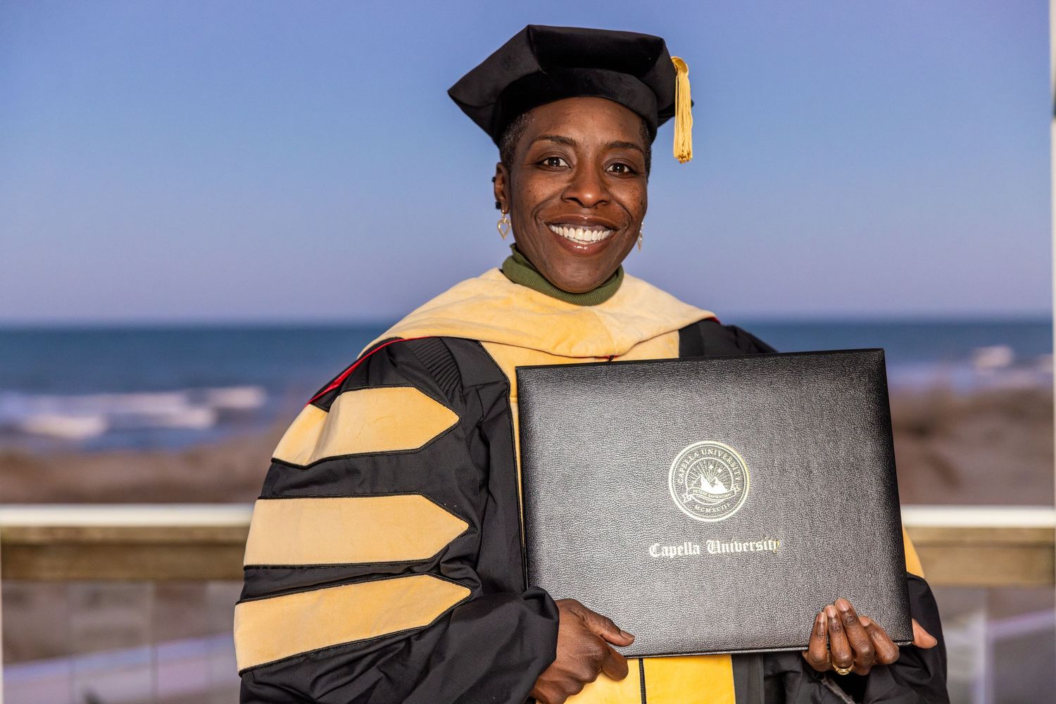 Woman in doctoral graduation regalia smiles holding a Capella University diploma by the ocean.