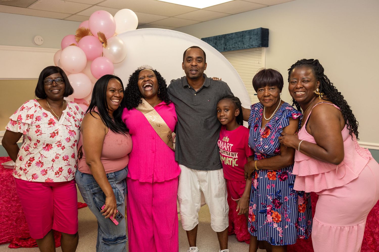 Group of people dressed in pink celebrating together at a party with balloons in the background.