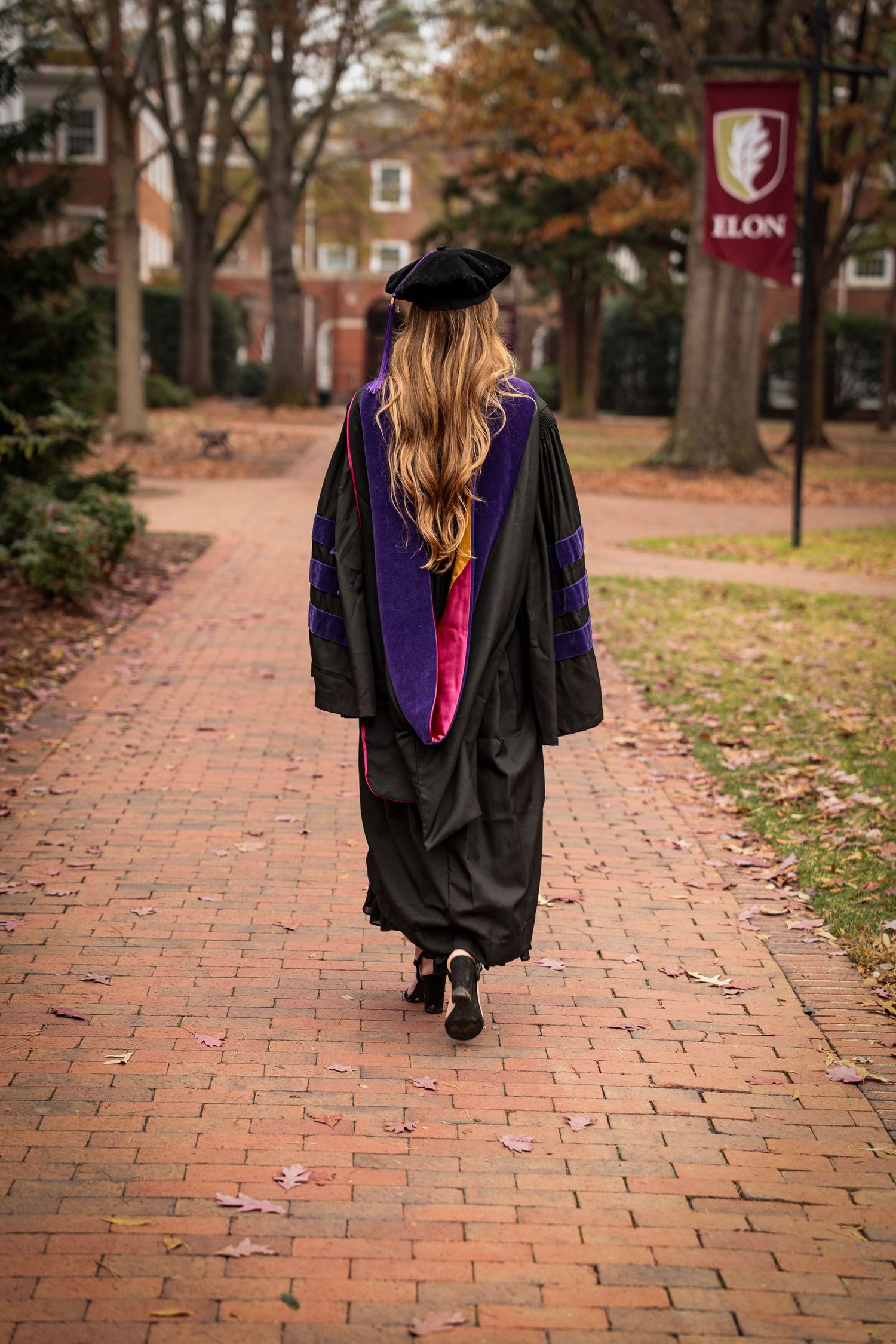 Graduate in academic regalia walking away on brick pathway with fall foliage.