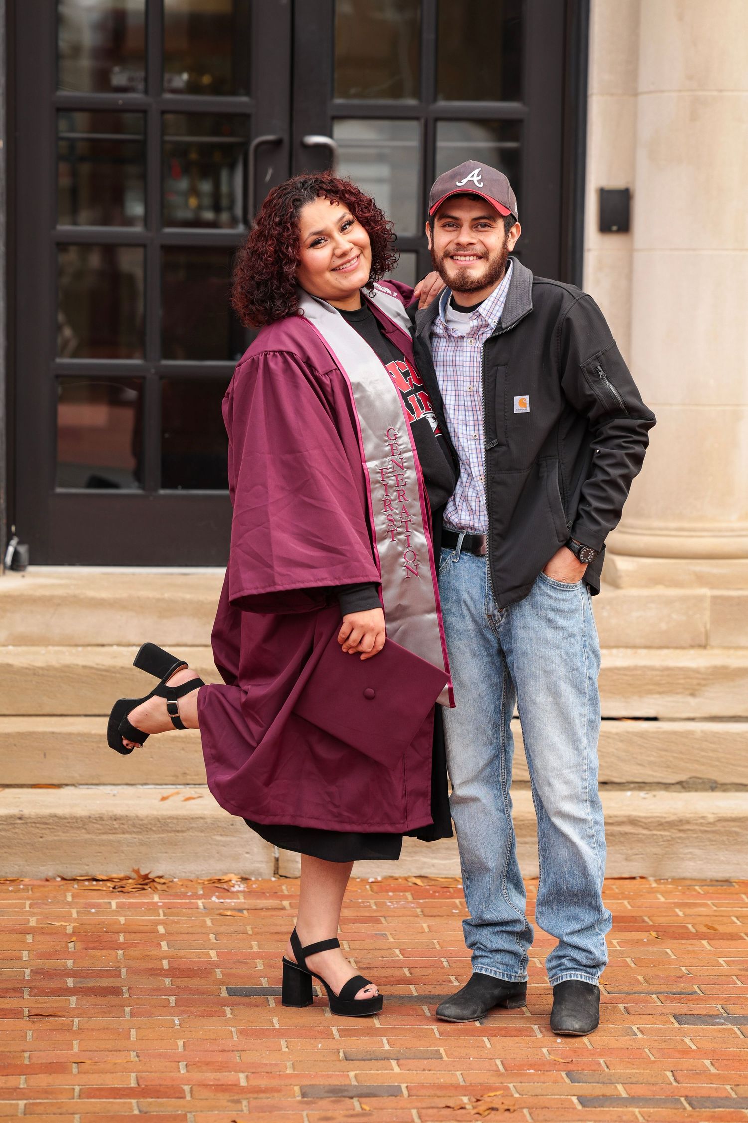 A couple stands together on red brick pavement in front of an administrative building entrance.