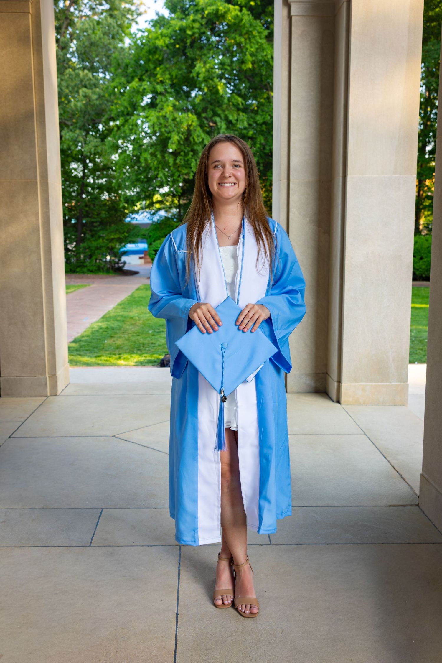 Young woman smiling in light blue graduation cap and gown, standing between stone columns with green trees in background.