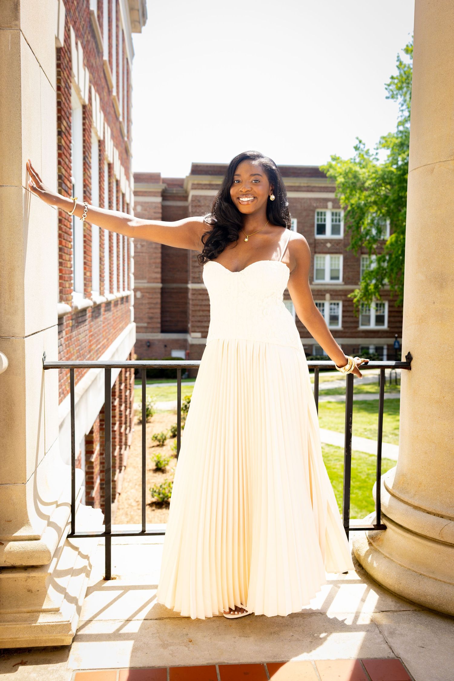 Young Black woman in elegant cream pleated gown smiling on a brick building balcony on a sunny day.