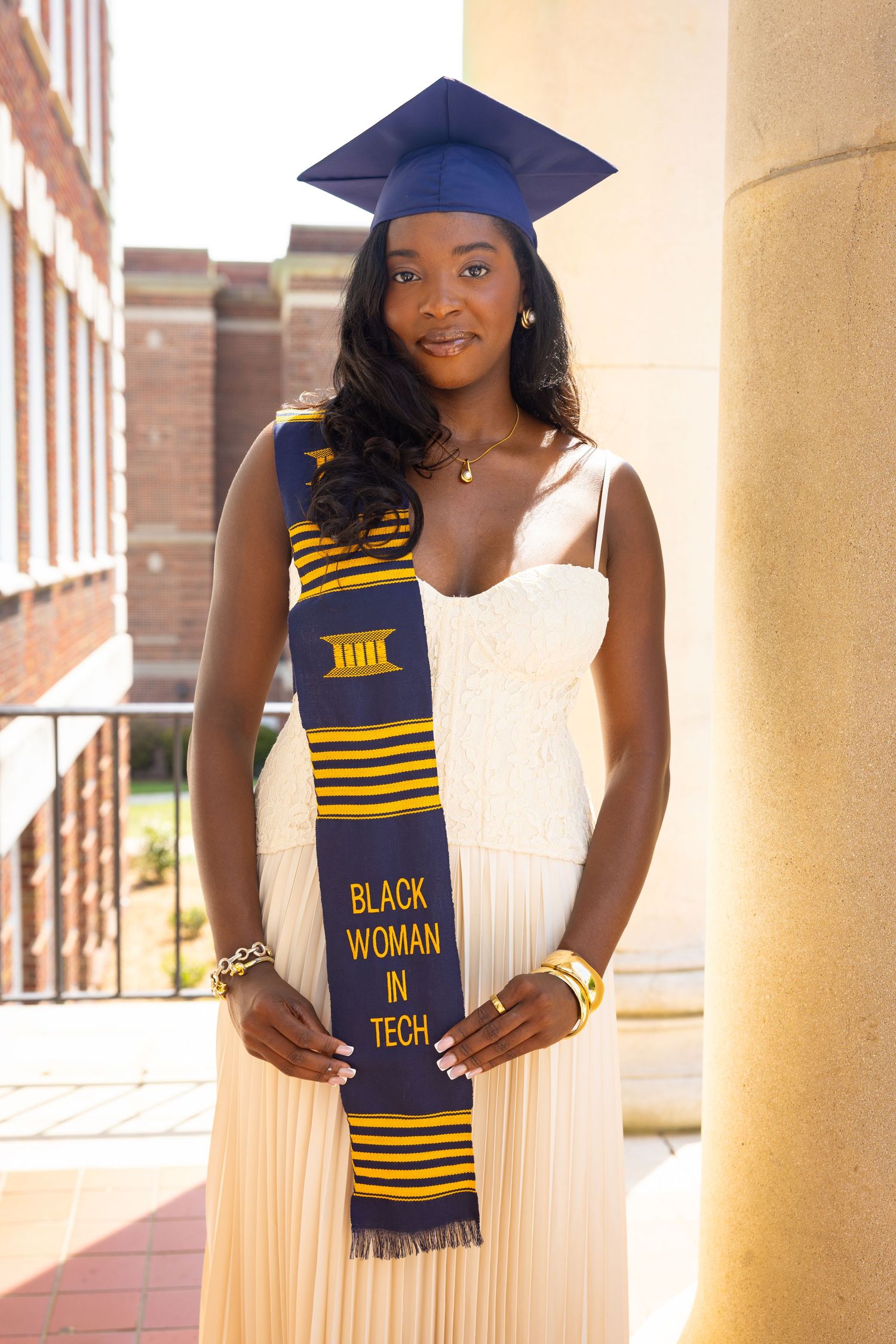 Graduate in cap and gown wearing a Black Woman in Tech sash poses proudly by a column on campus.