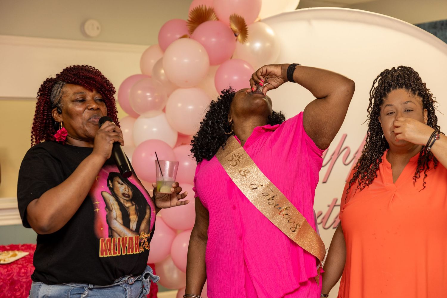 Three women celebrate a bachelorette party with pink balloons, one wearing a bride-to-be sash at an indoor event.