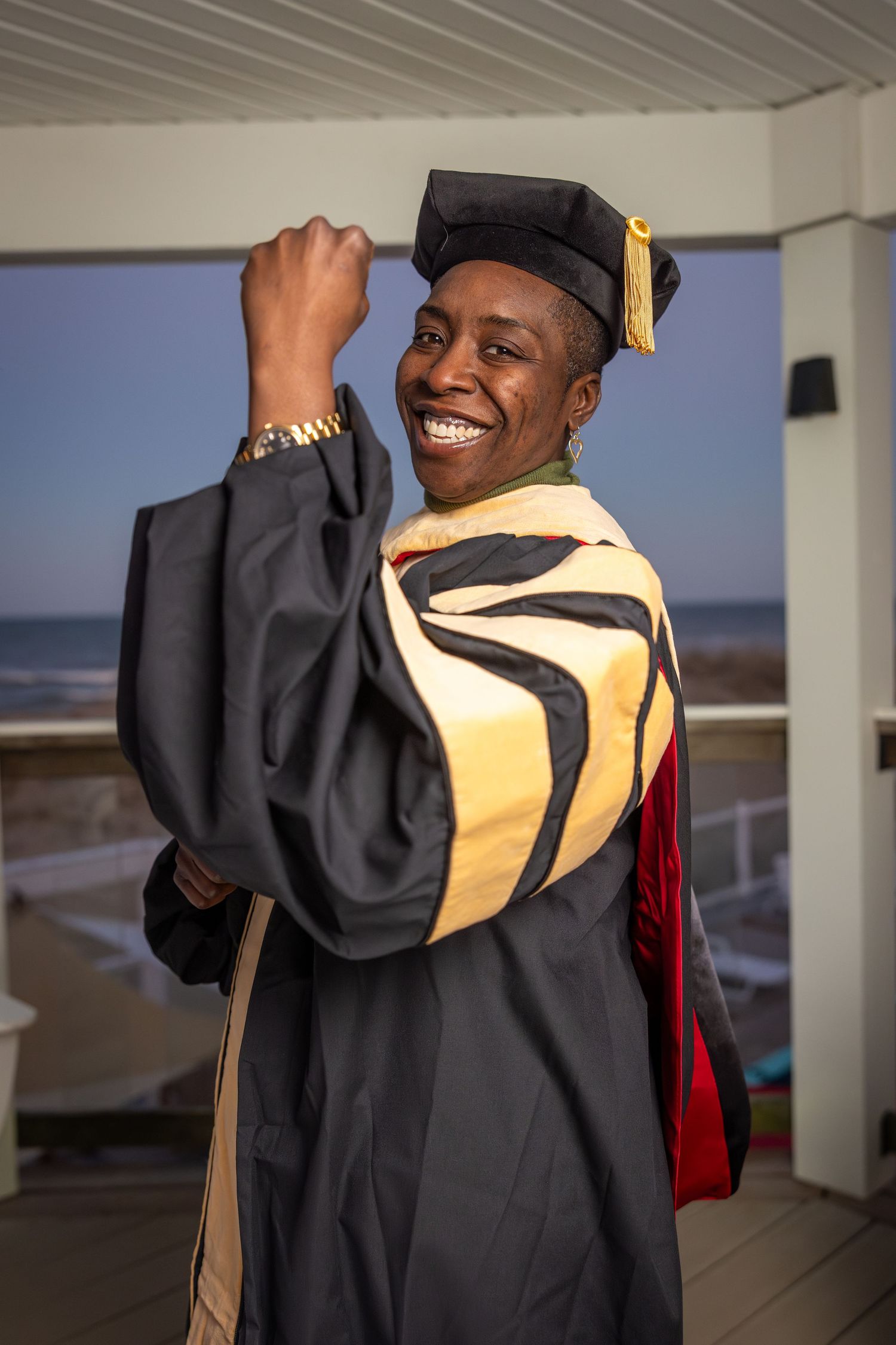 Smiling graduate in black cap and gown with gold and red hood poses outdoors near beach at sunset.