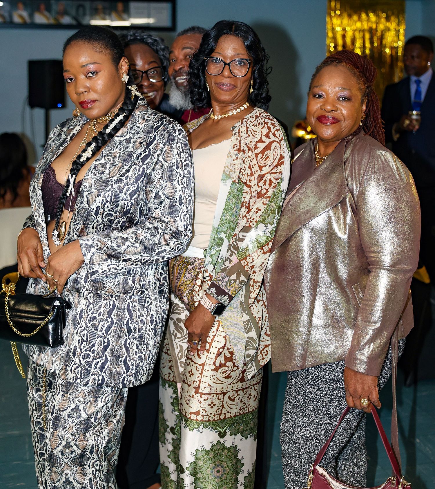 Guests in glamorous evening wear including sparkly dresses and metallic outfits pose together at a formal celebration event.
