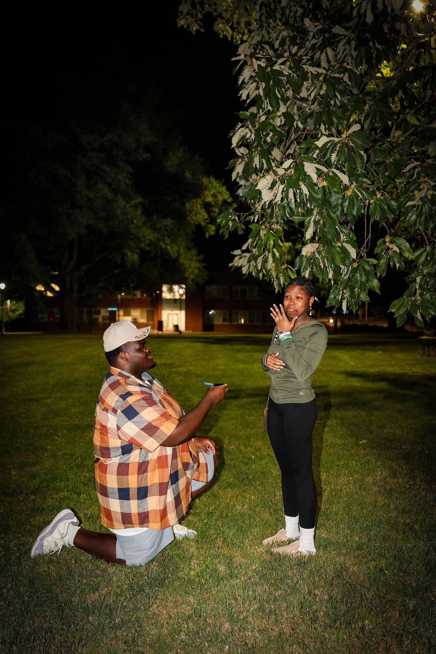 A romantic proposal scene under trees at night with campus buildings in the distance.