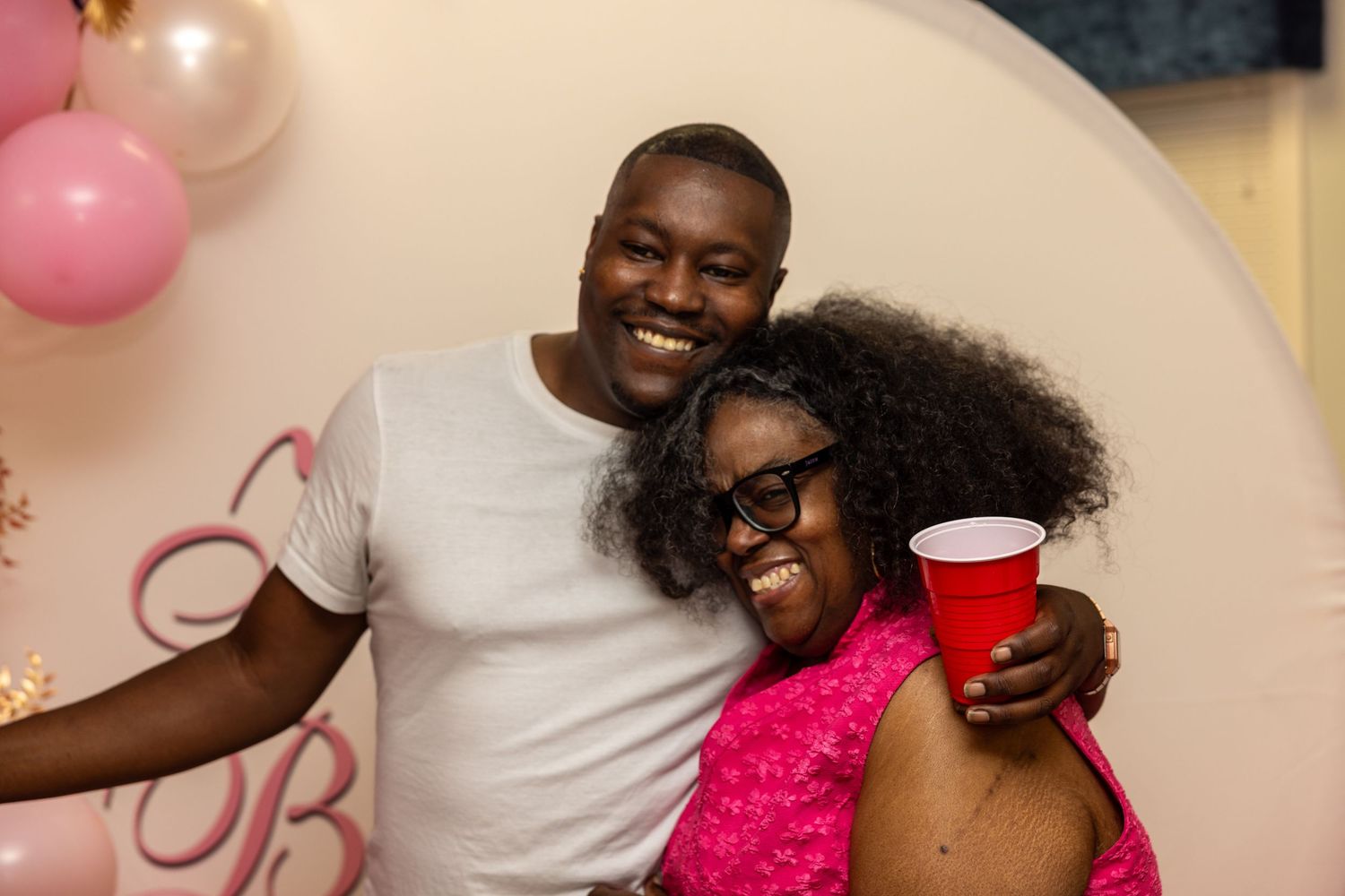 A smiling man and woman in pink dress hugging and posing together at a pink-themed celebration party.