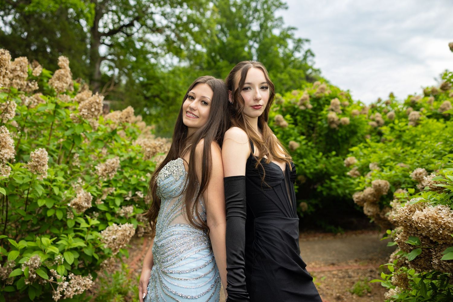 Two young women in formal dresses pose back-to-back in a lush garden with blooming hydrangeas.