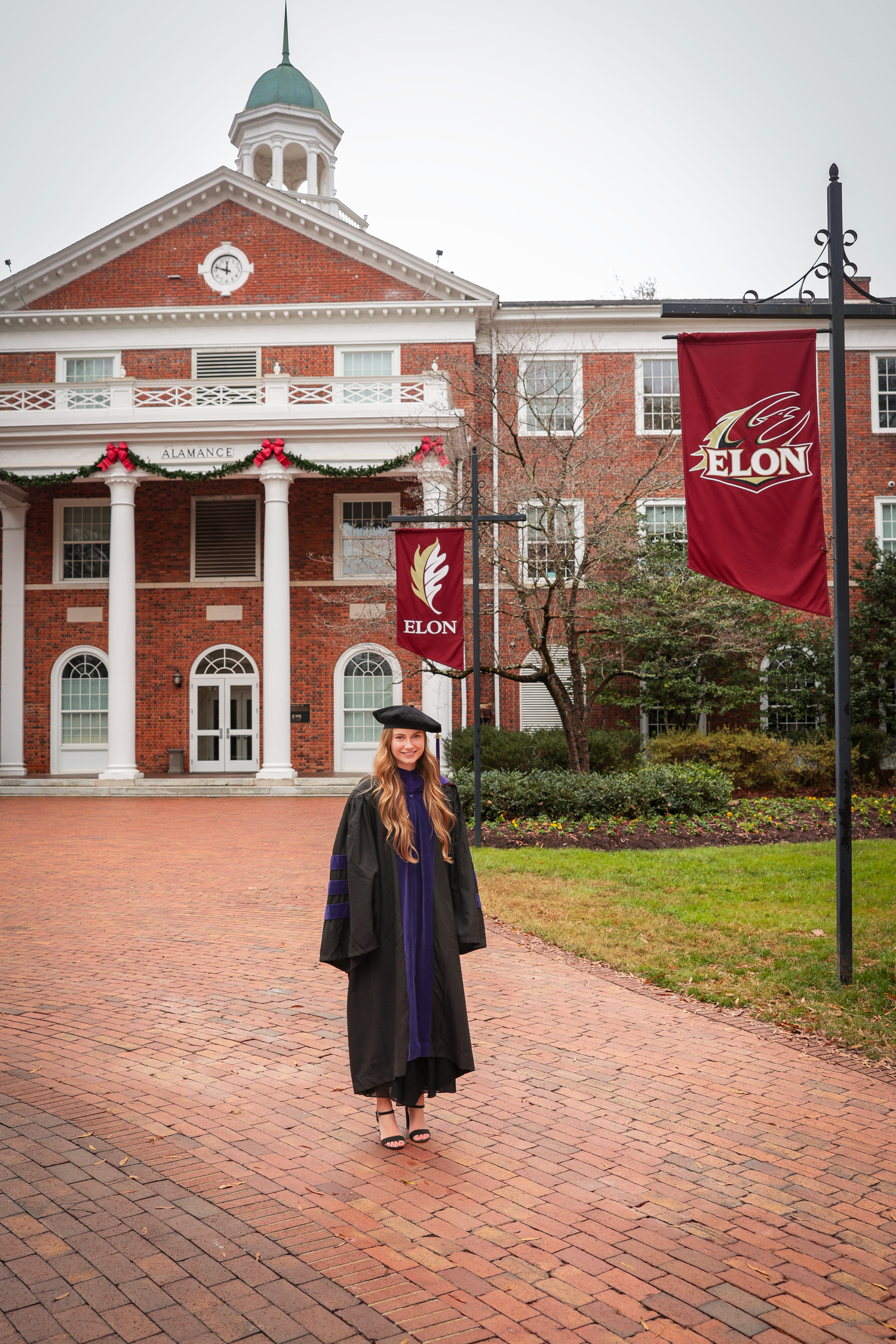Graduate in cap and gown stands with red banner in front of brick university building.