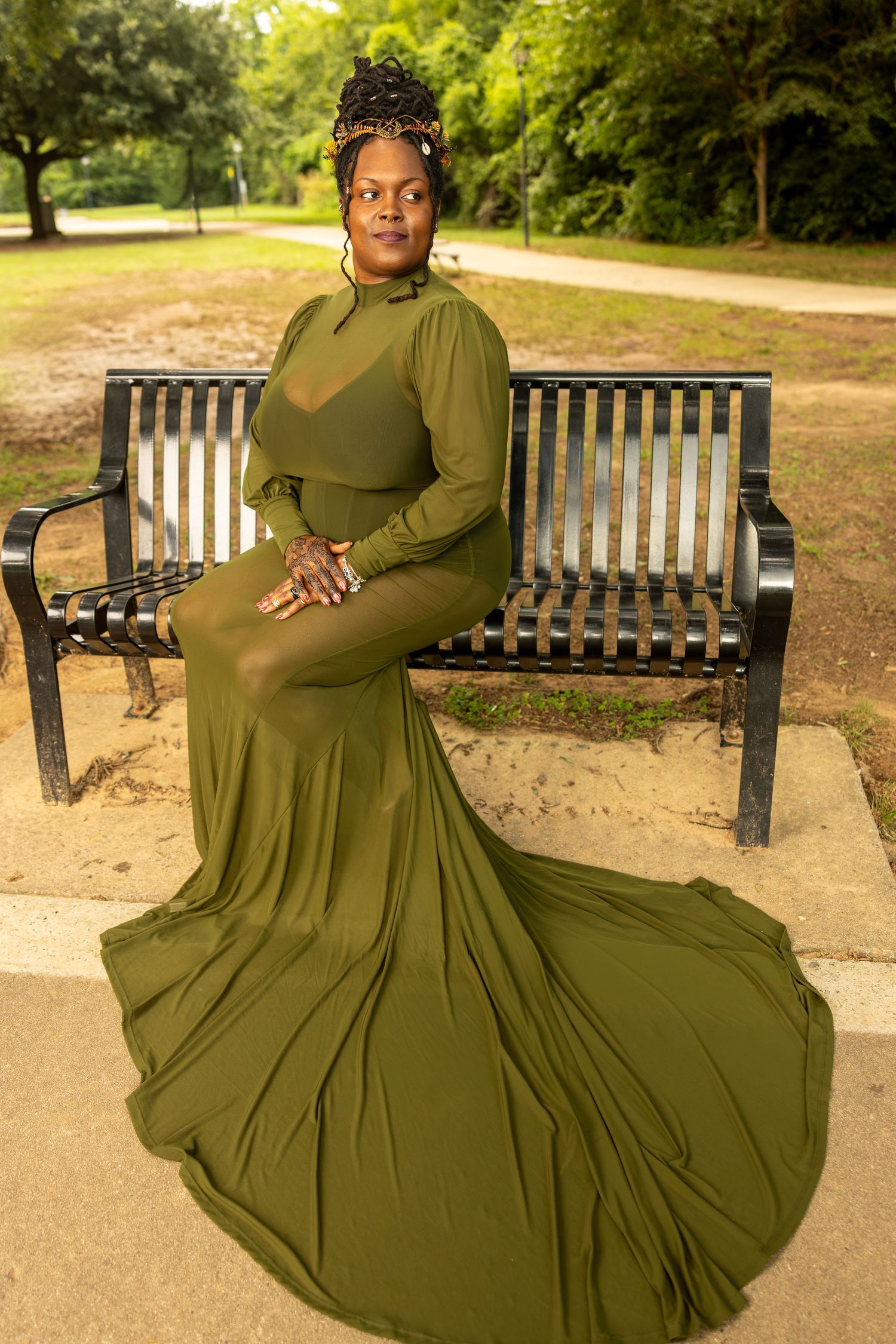 Woman in elegant olive green gown with long train sits gracefully on park bench outdoors.