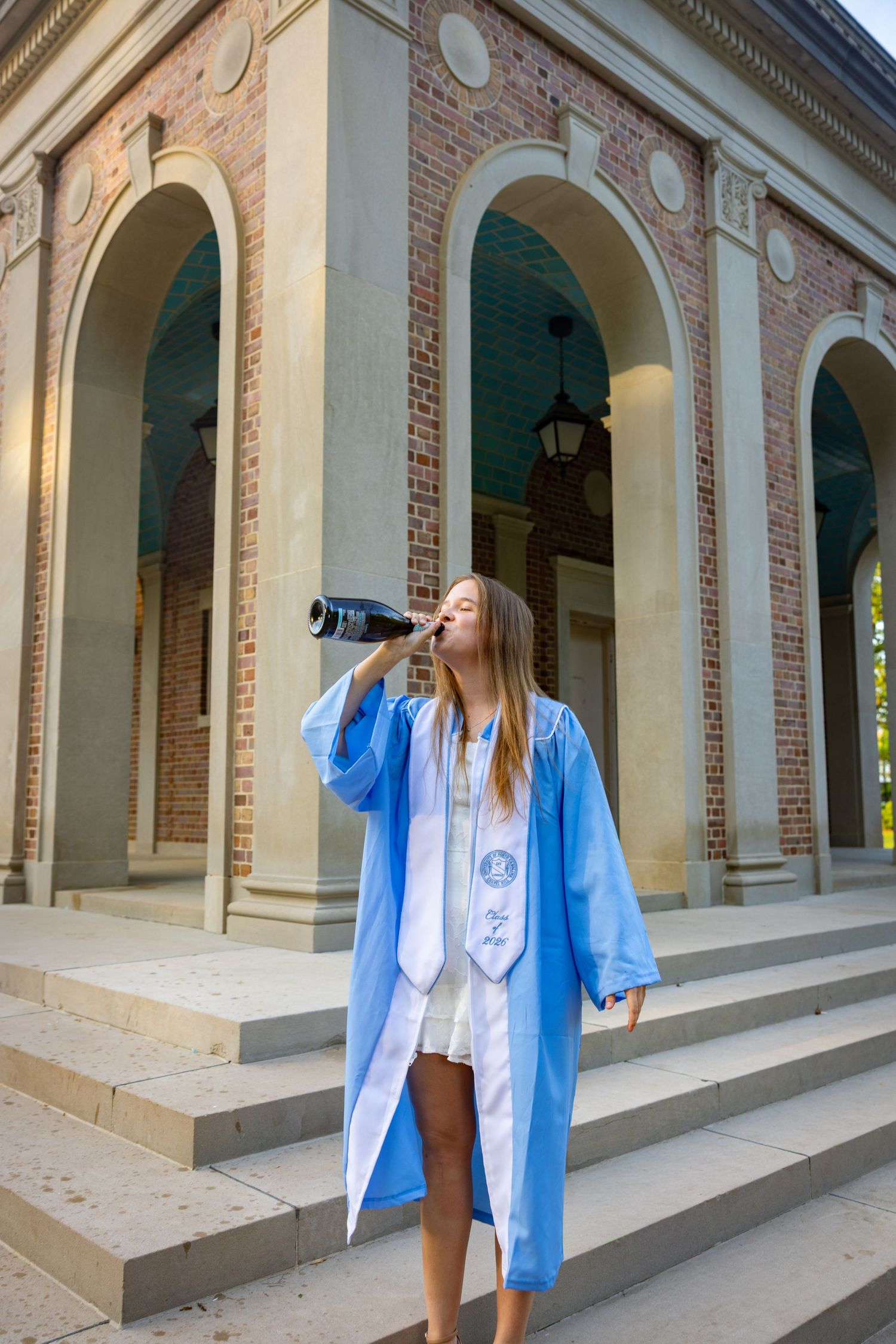 A young woman in a light blue graduation gown drinks from a bottle on steps outside a brick building with arched columns.