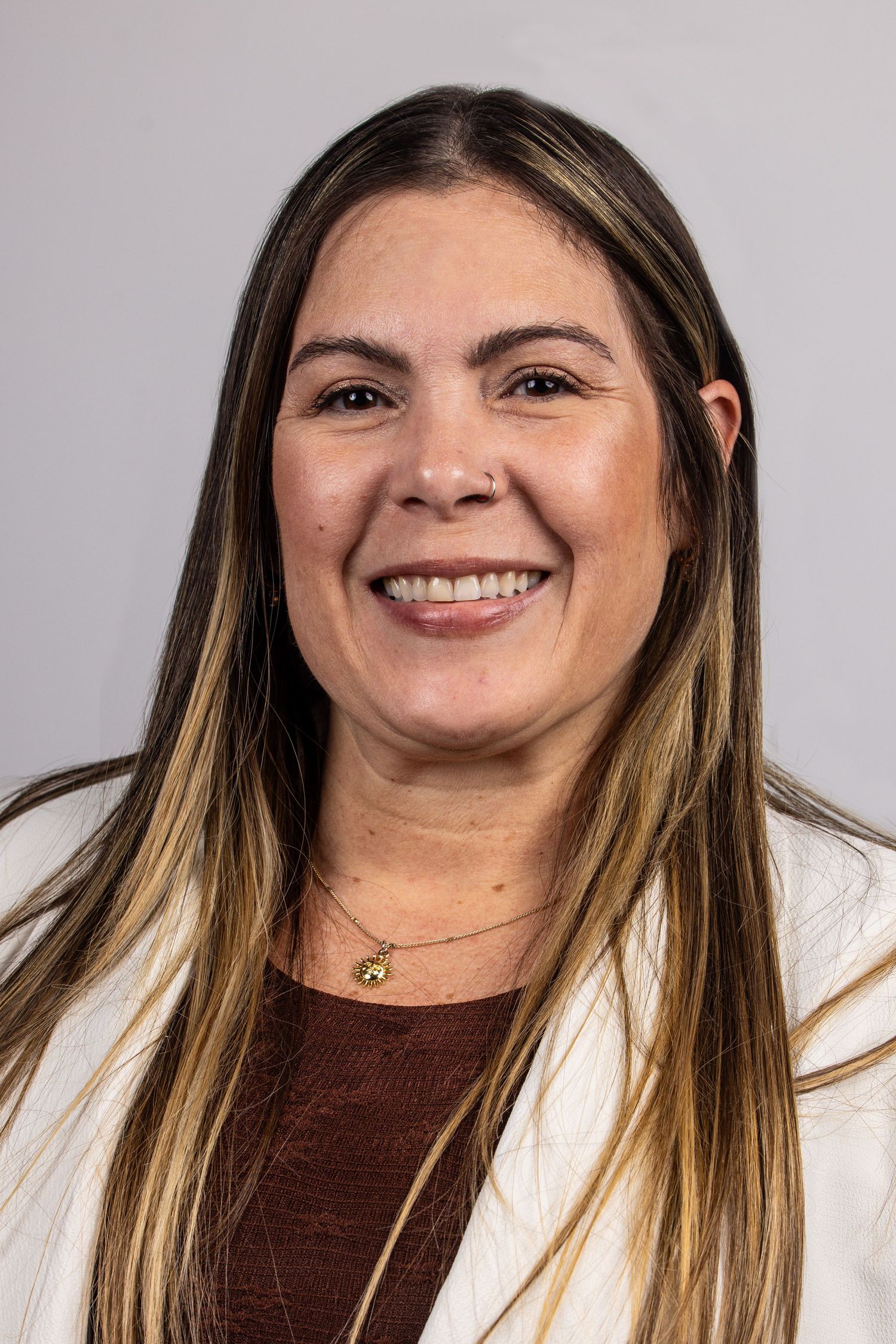 Smiling woman with long brown highlighted hair wearing a white blazer and burgundy top with a gold necklace.