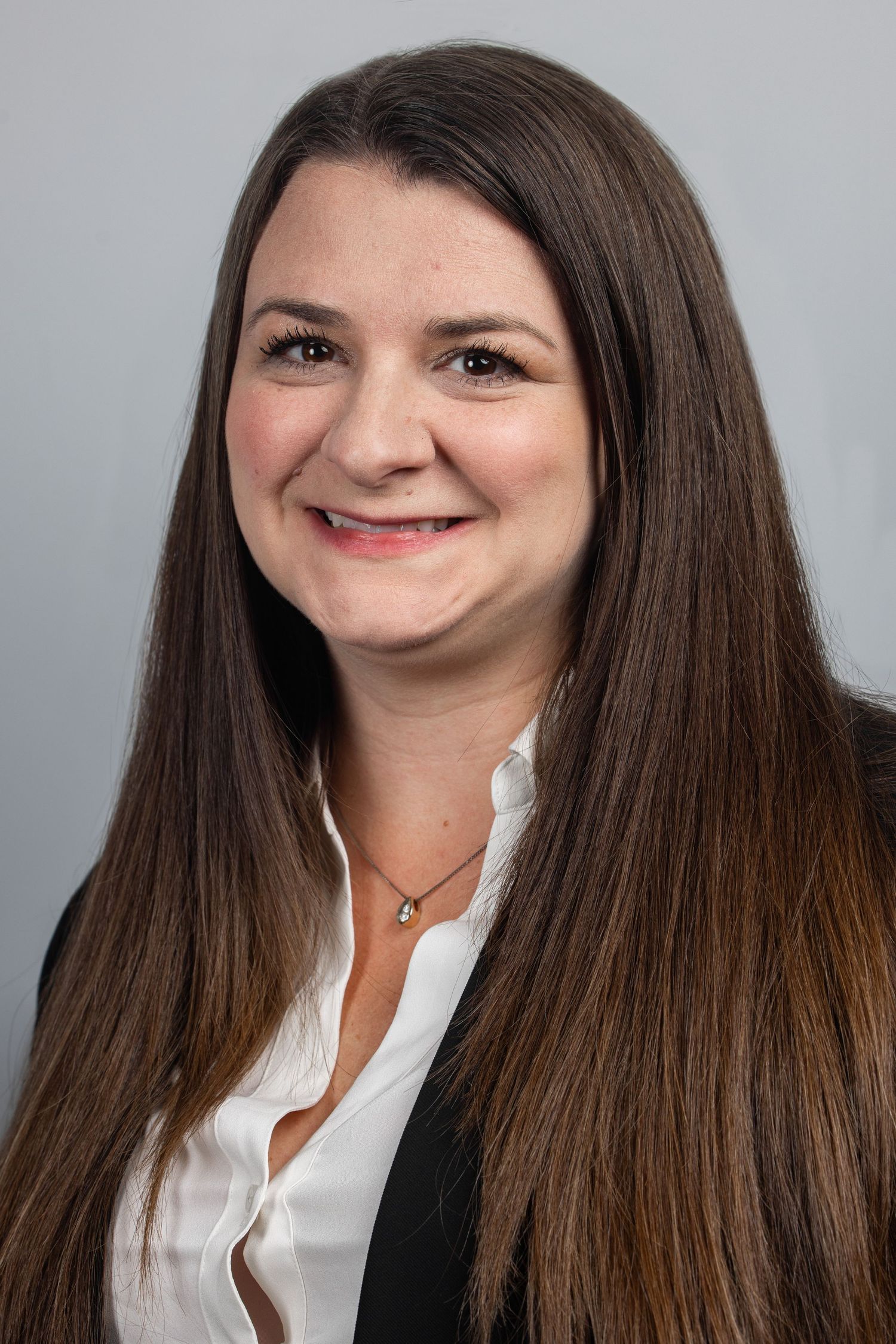 Professional headshot of a smiling woman with long brown hair wearing a white blouse and black blazer.