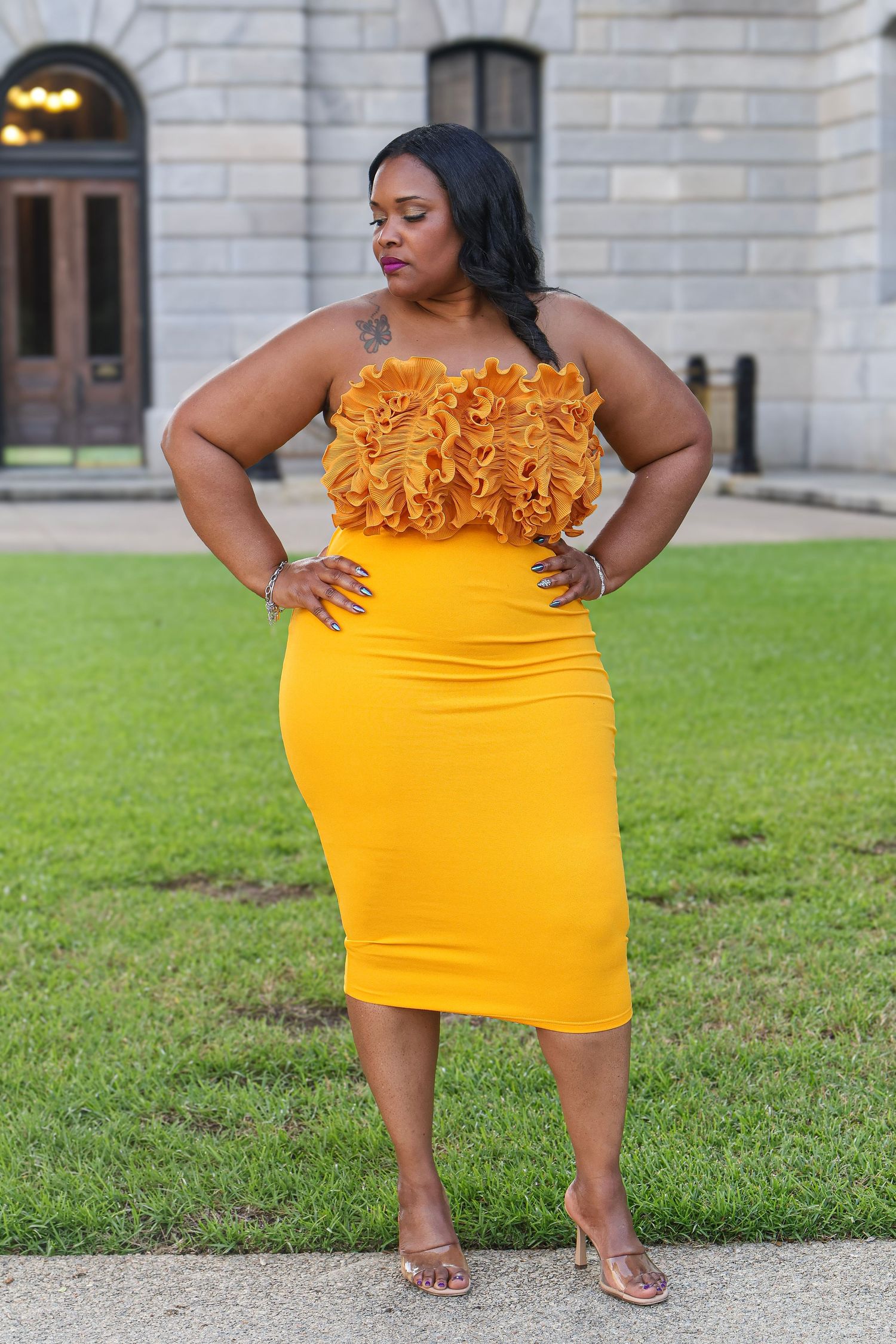 A bright yellow bodycon dress with ruffled neckline detail photographed against a stone building exterior.