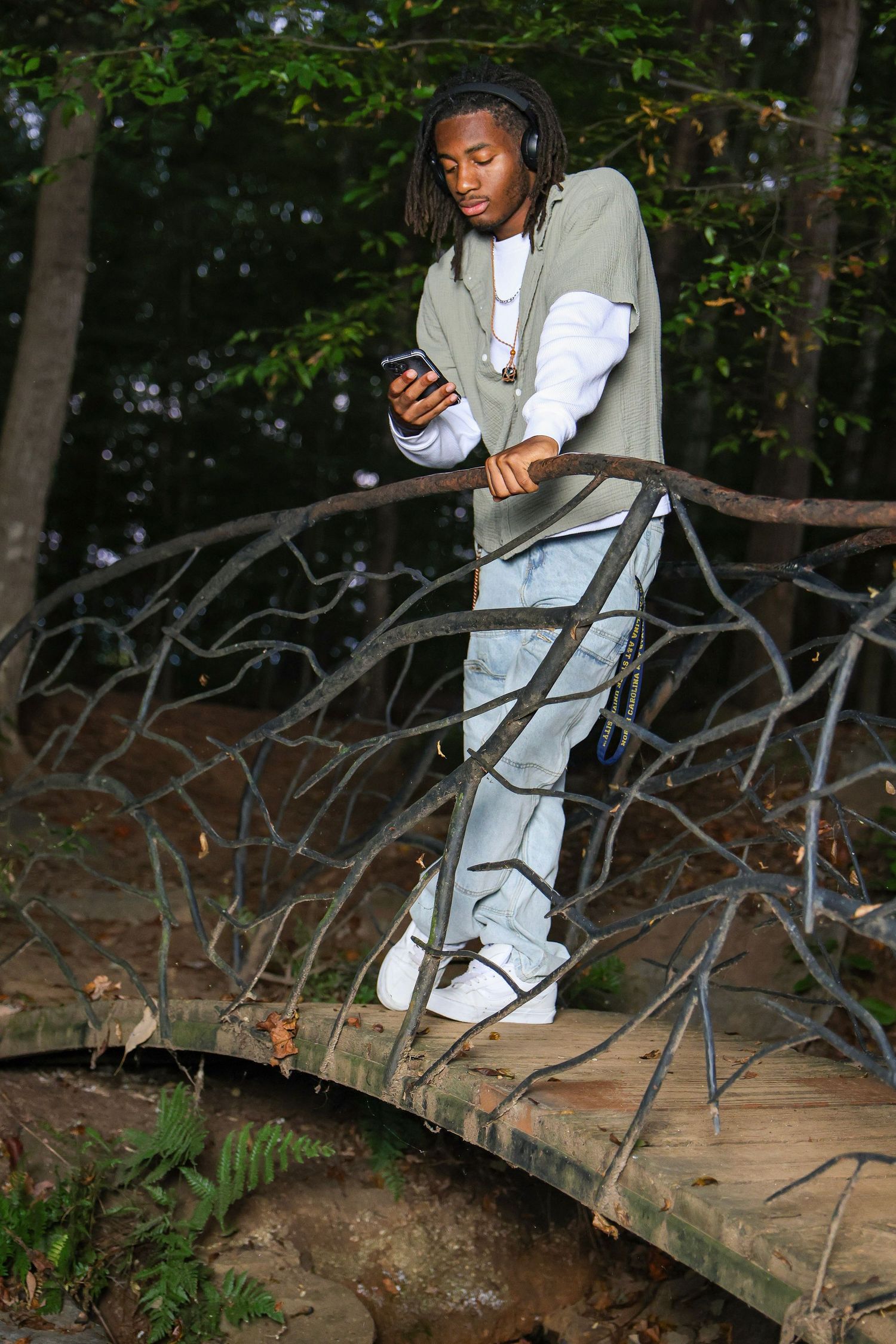 Person sitting on fallen tree branch in casual attire while using phone in wooded area.