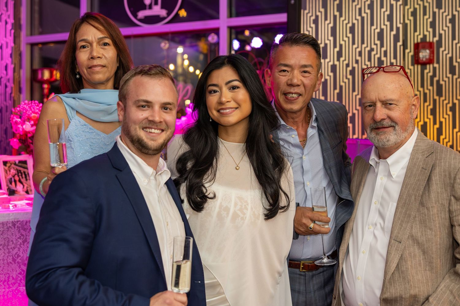 Five guests smiling and posing together at an elegant pink-lit event, one holding a champagne glass.