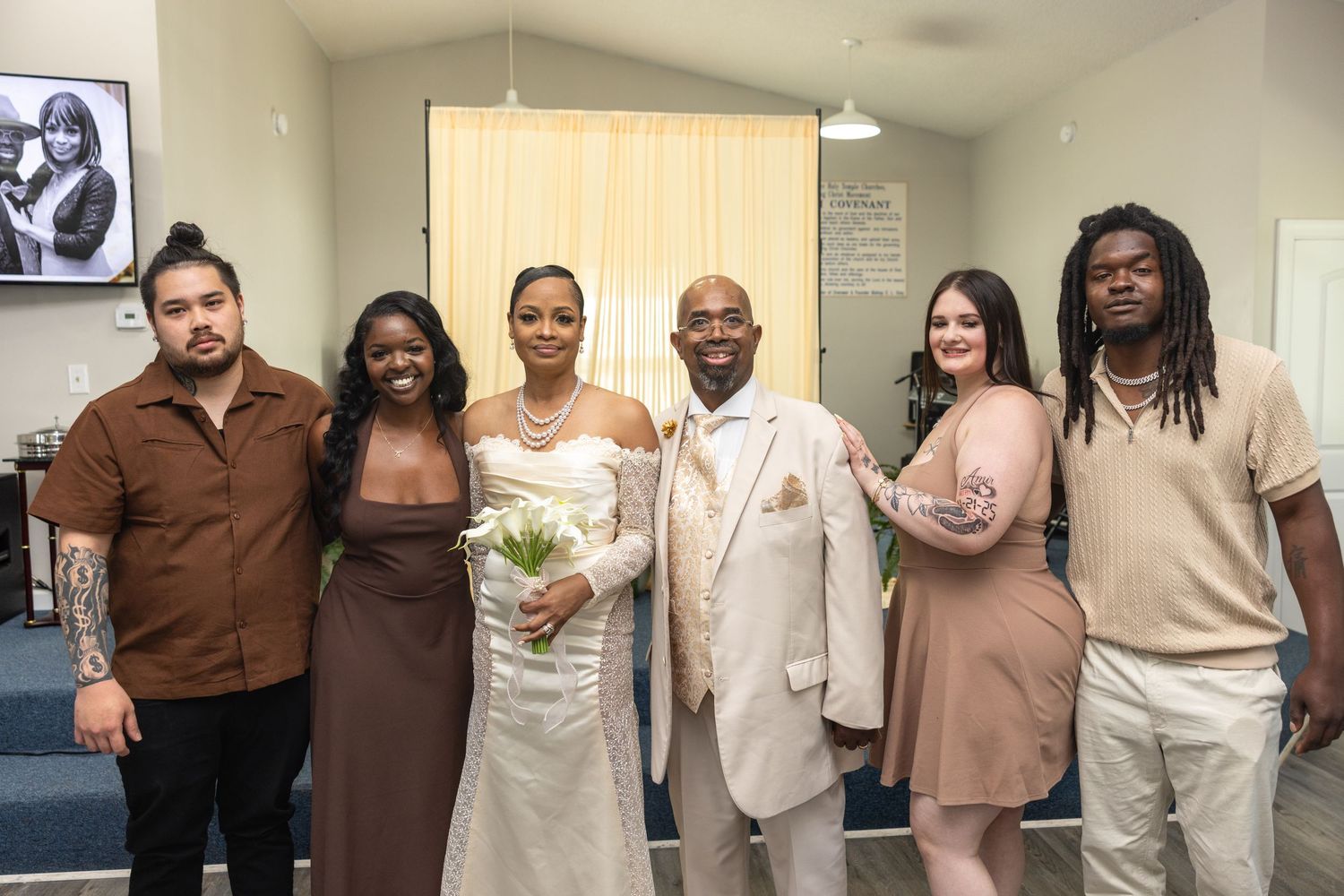 A joyful wedding party of six poses together in a small chapel, with the bride and groom centered among smiling family members.